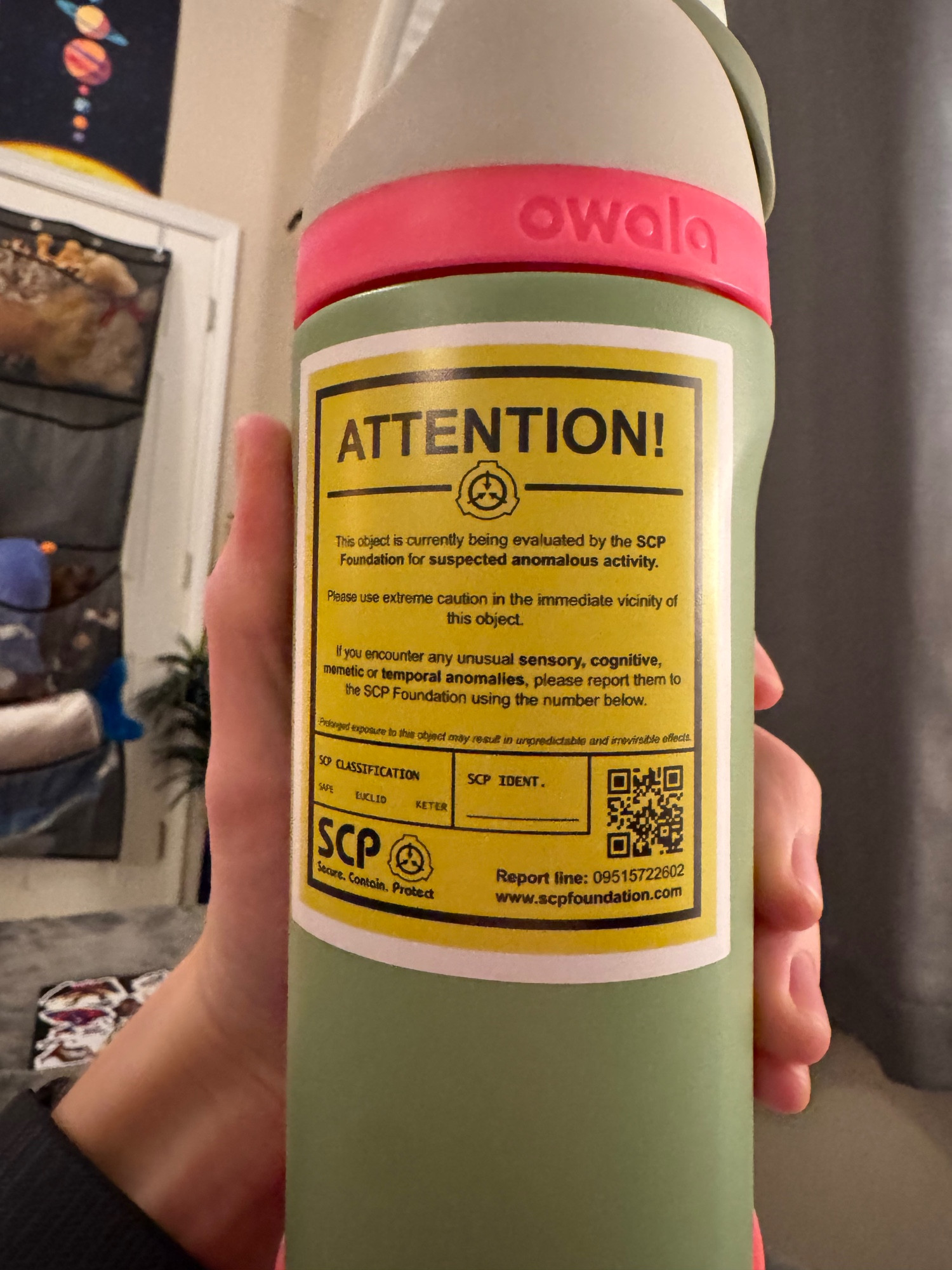 A yellow sticker on a mint green water bottle that says "ATTENTION! This object is currently being evaluated by the SCP Foundation for suspected anomalous activity. Please use extreme caution in the immediate vicinity of this object. If you encounter any unusual sensory, cognitive, memetic, or temporal anomalies, please report them to the SCP Foundation using the number below." There is a fillable box that is labeled "SCP CLASSIFICATION" with the options SAFE, EUCLID, and KETER. There is another box labeled "SCP IDENT." with a space to fill out a number. There is a QR code in the bottom right corner of the sticker that leads to the SCP wiki when scanned. Bellow that code is a phone number labeled "Report line" and a website address that reads "www.scpfoundation.com." In the bottom left corner is the SCP logo with the text "Secure. Contain. Protect." bellow it.