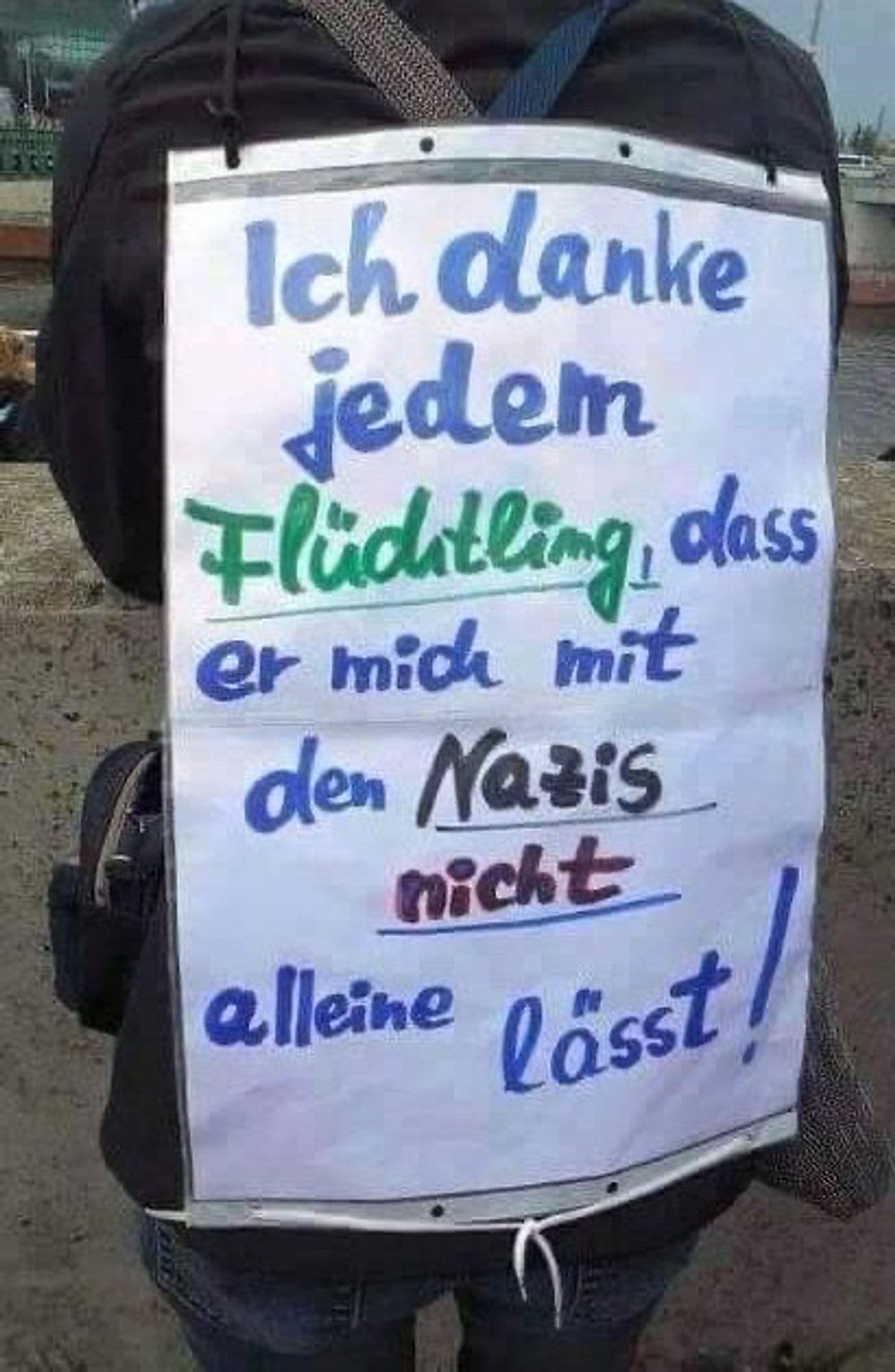 Ein Mensch, der sich mit in Plakat auf den Rücken geschnallt hat auf dem steht:
„Ich danke jedem Flüchtling, dass er mich mit den Nazis nicht alleine lässt.“