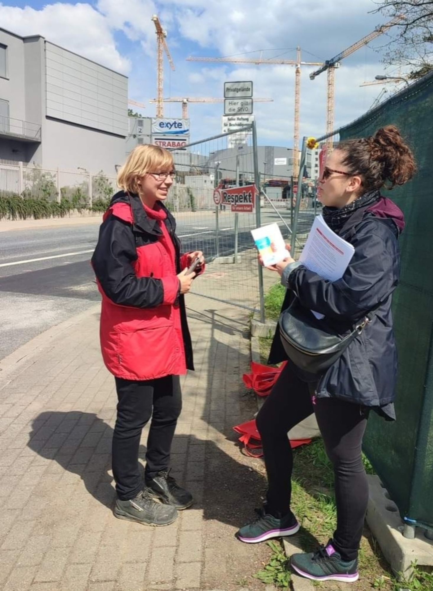 Auf dem Bild ist im Hintergrund eine Großbaustelle zu sehen. Im Vordergrund zwei Frauen mit Infomaterialien.