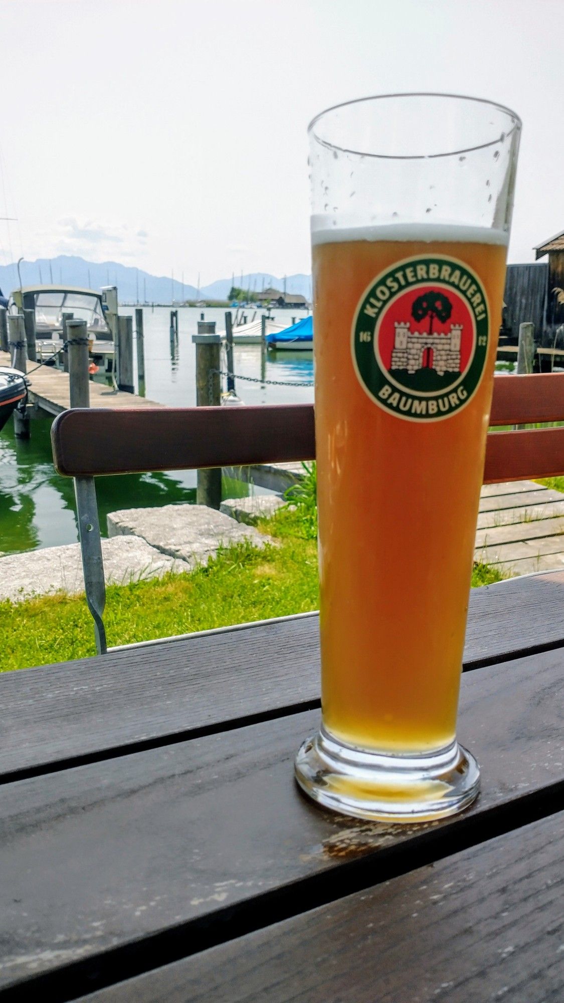 Glas Weißbier auf einem Tisch, dahinter Steg und Boote, Chiemsee und Berge.