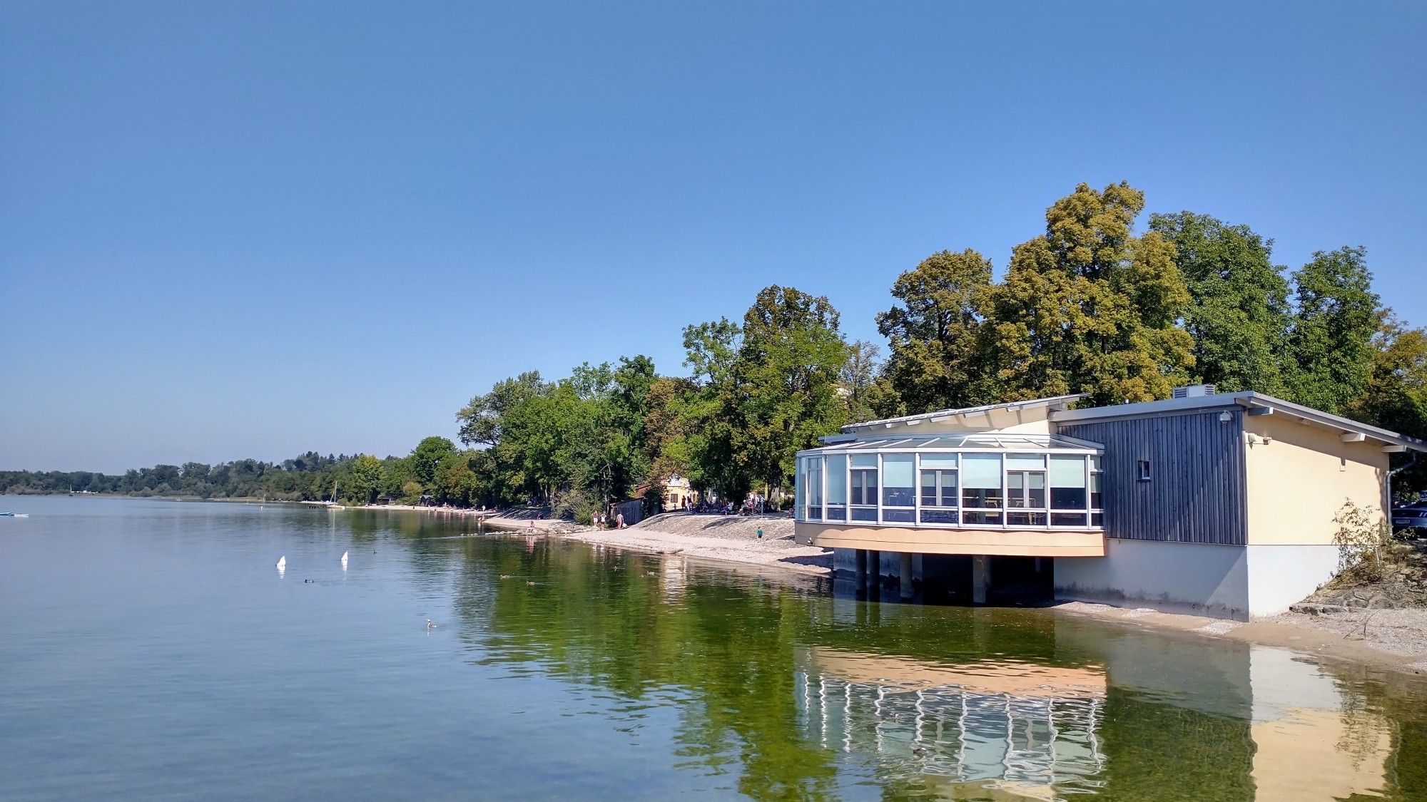 Blick auf das Chiemsee-Ufer in Chieming. Im Wasser spiegelt sich ein Haus und Bäume.