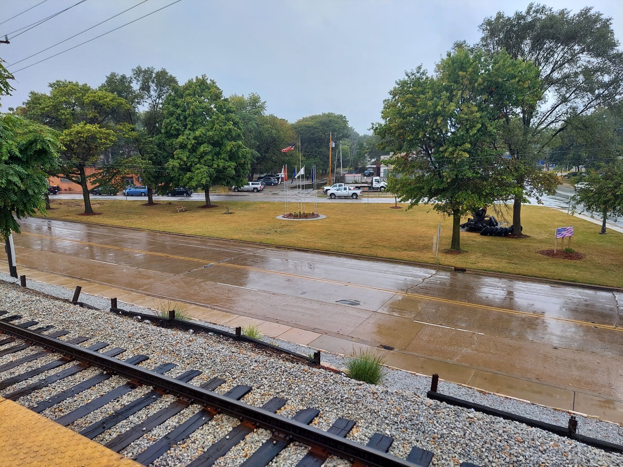 Foreground: train track, lower left. Street wet with rain. Park with brown grass, trees on left and right ends. Circle with flagpoles in center of Park. American Legion hall beyond trees on left.