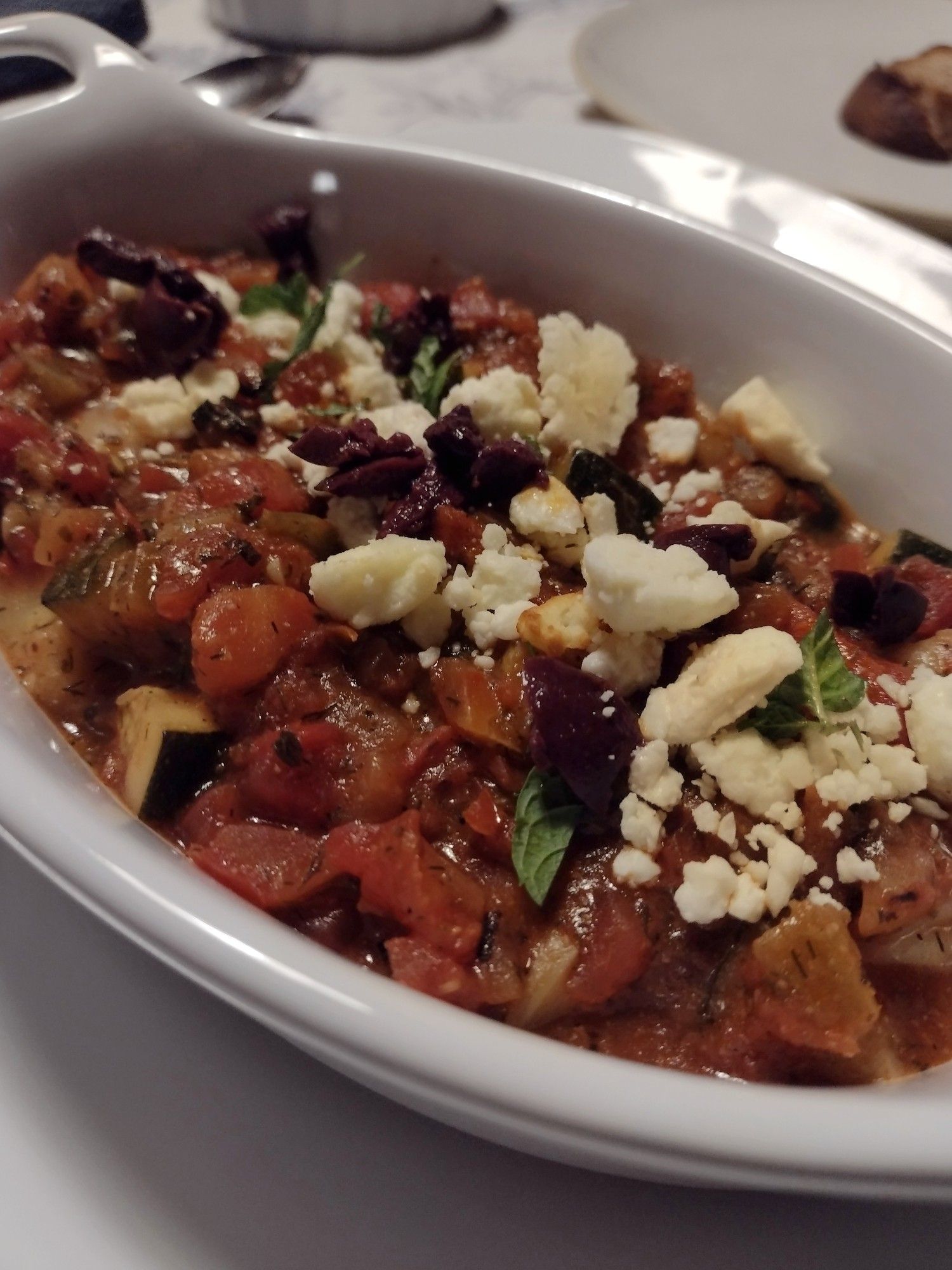 A close-up image of a dish of Shrimp Saganaki, featuring a rich tomato sauce with chunks of tomatoes and herbs. The sauce is garnished with crumbled feta cheese and sliced black olives, adding a contrast of white and dark colors to the red sauce. Fresh green herbs are sprinkled on top for added color and flavor. The shrimp is not visible as it is covered by the sauce.