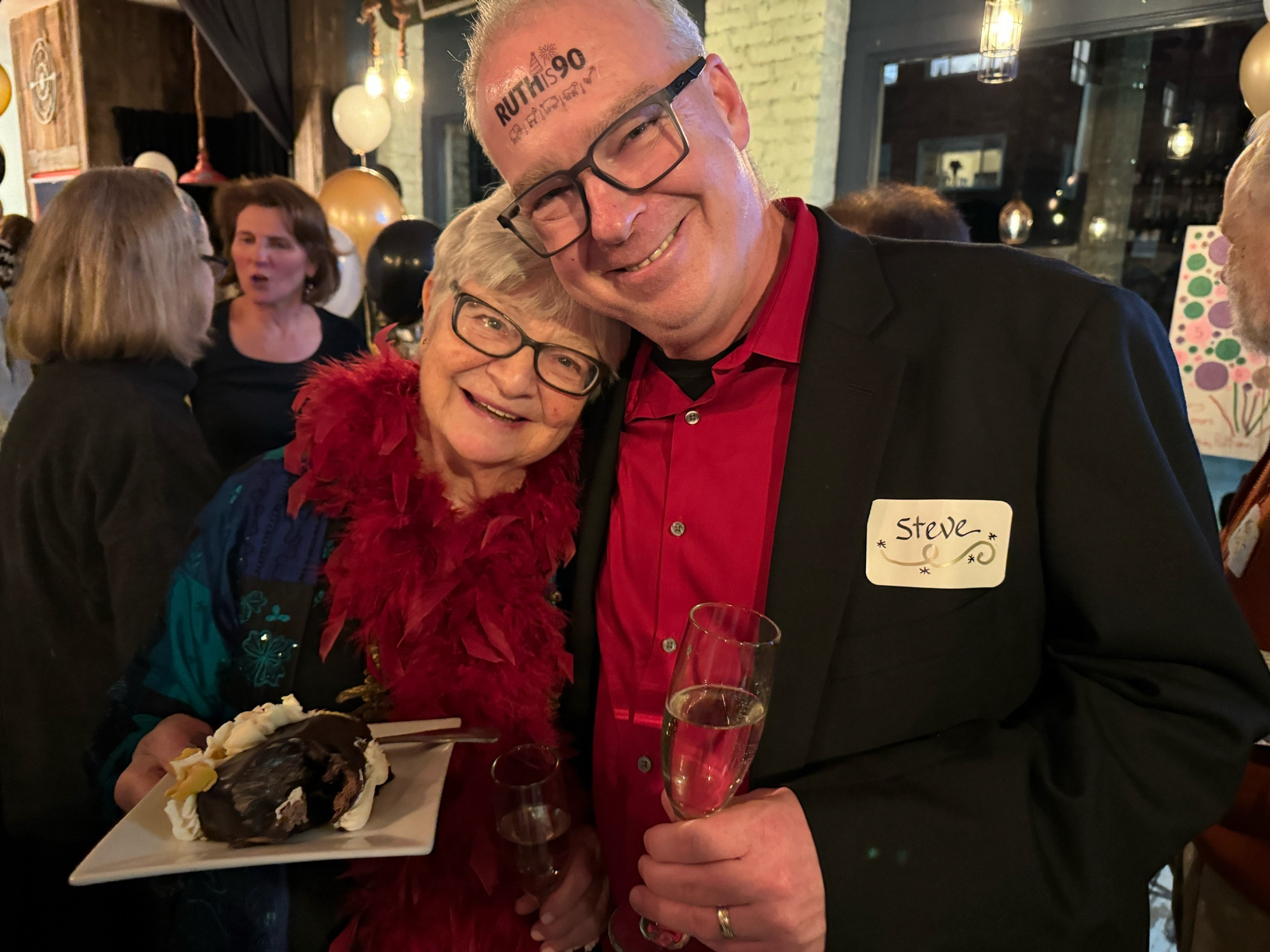 Photo shows a celebrating Steve with a temporary forehead tattoo reading “Ruth is 90” all smiles and arm in arm with the fabulous Ruth herself.