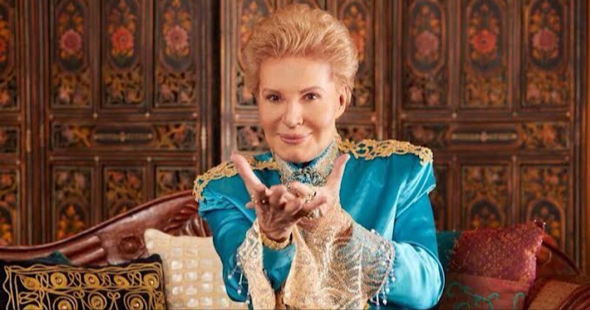 Walter Mercado in a turquoise and gold jacket. He has his hands up in a blessing motion. He was truly magic