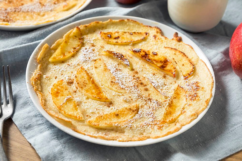 A photo of a plated crêpe with apple slices baked into it, with powdered sugar sprinkled on top.