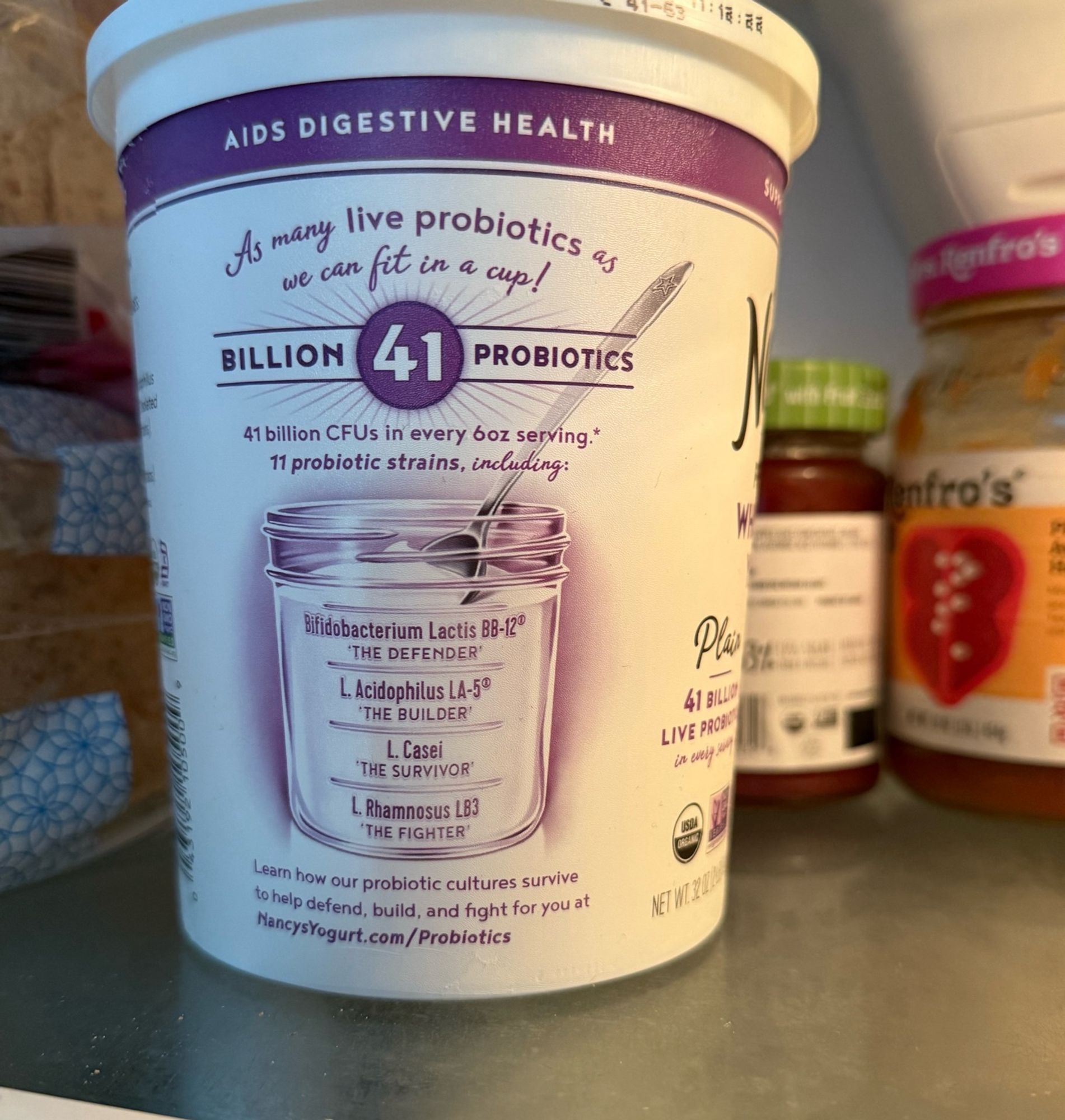 A pint yogurt container in a refrigerator. It lists several kinds of cultures on the back label, labeled in sequence, the Defender, the Builder, the Survivor, and the Fighter.