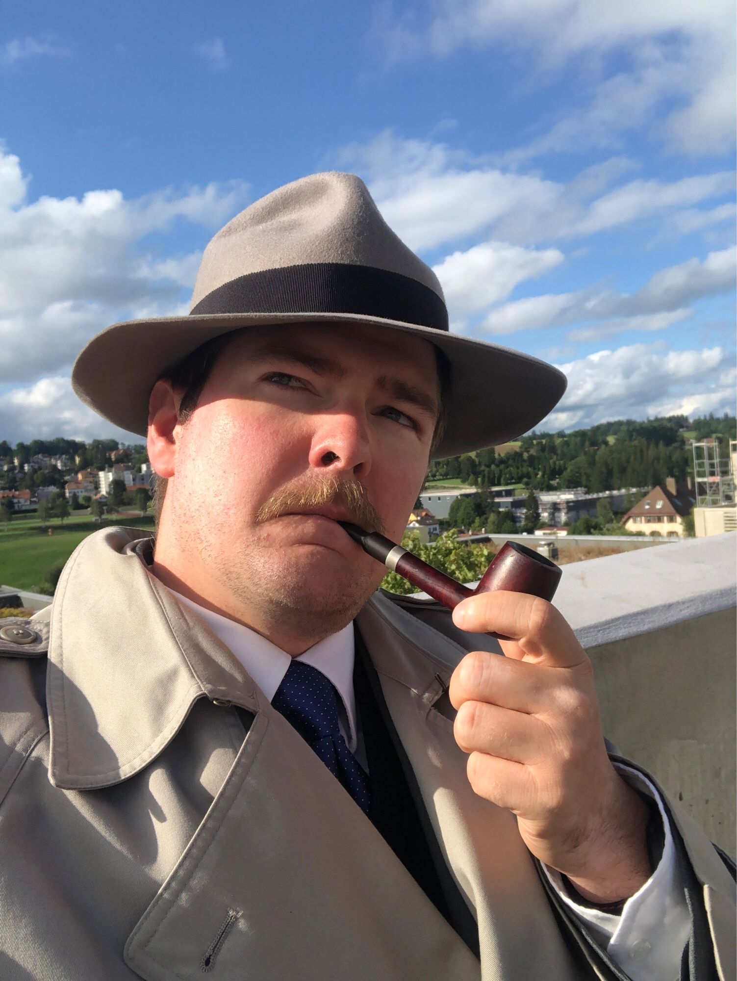 Selfie showing a 30 something man with a hat, trench-coat and pipe, looking up, a small Swiss town in the background