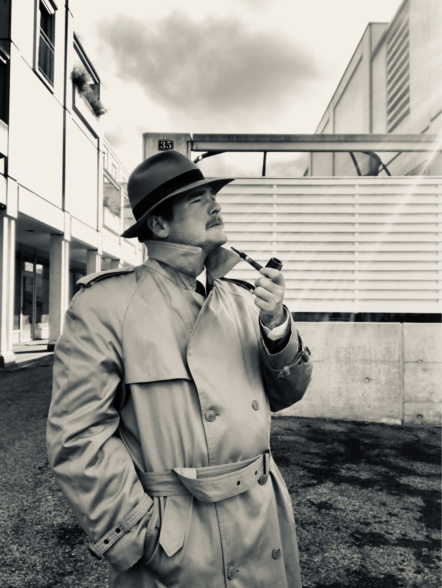 Black and white digital photography of the author as Chief-Inspector Maigret, wearing a trench-coat, soft hat, holding a pipe in his left hand, looking up wistfully. It takes place in an urban complex in La Chaux-de-Fonds, Switzerland, 2024