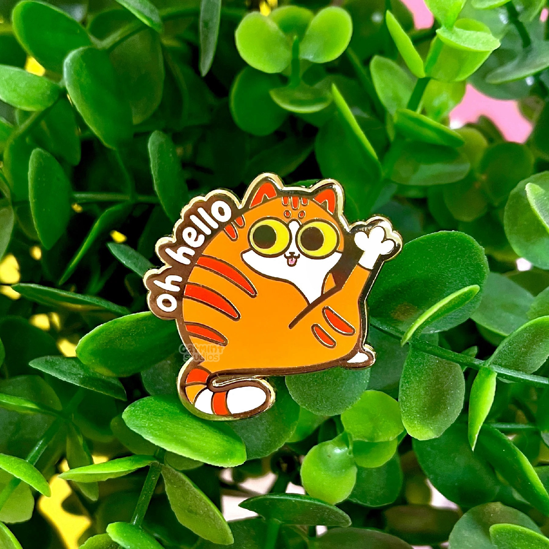 An enamel pin sits on a background of leaves. The design is of a chubby orange tabby cat with his tongue out and leg sticking up. The words “oh hello” are written above his back.