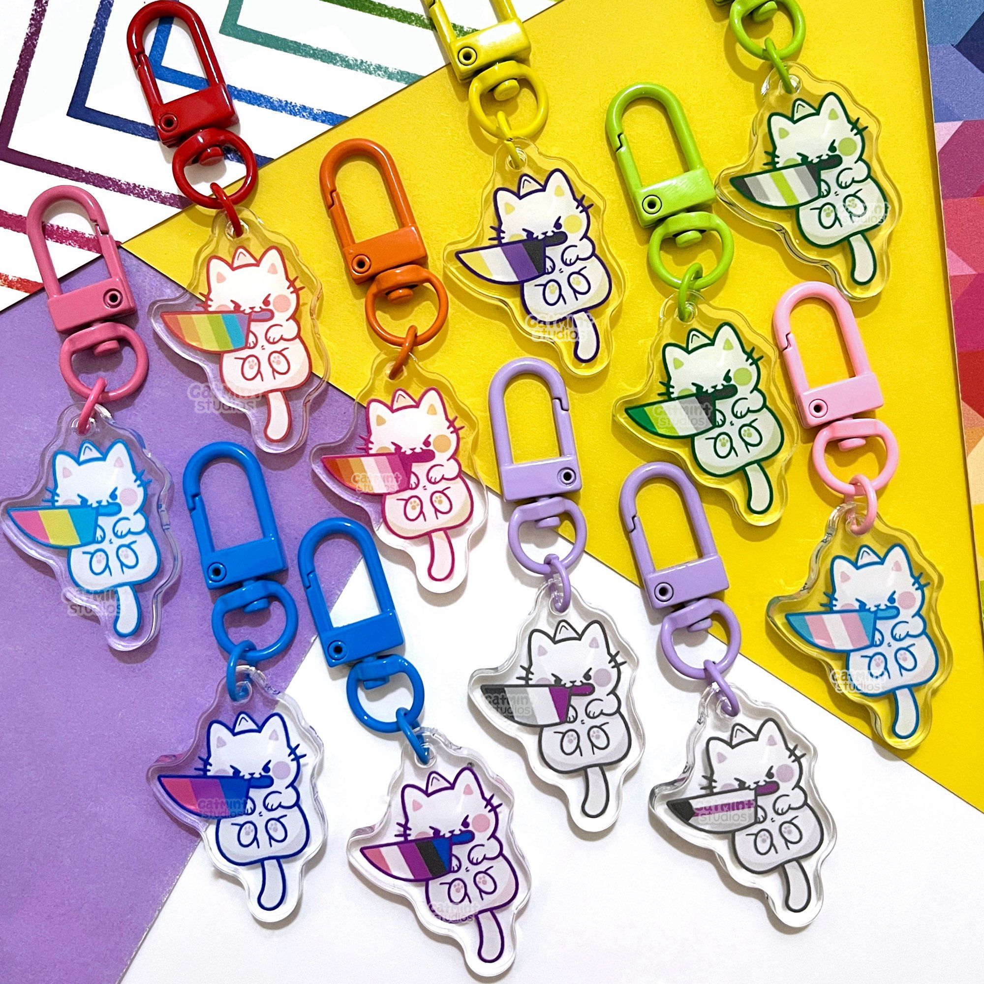 A collection of 11 small keychain charms sits on a purple, white and yellow background. The charm designs are all small cute white cats with angry eyes and knives in their mouths. All of the blades have different pride flag designs inside. All of the charms have different flags and include: gay, lesbian, pan, bi, genderfluid, ace, demi, trans, aro, agender, and non-binary.