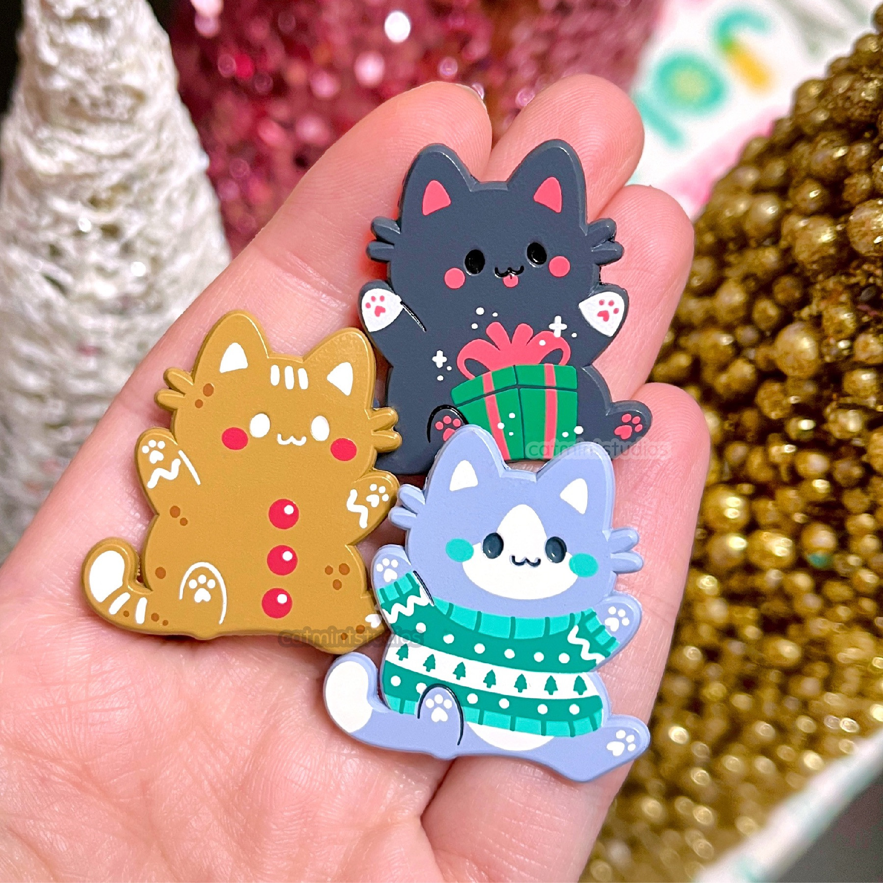 A hand holding three enamel pins of cats. One looks like a gingerbread cookie, one is a black cat holding a small gift between its paws, and one is a gray & white cat with a teal & white striped ugly Christmas sweater on.