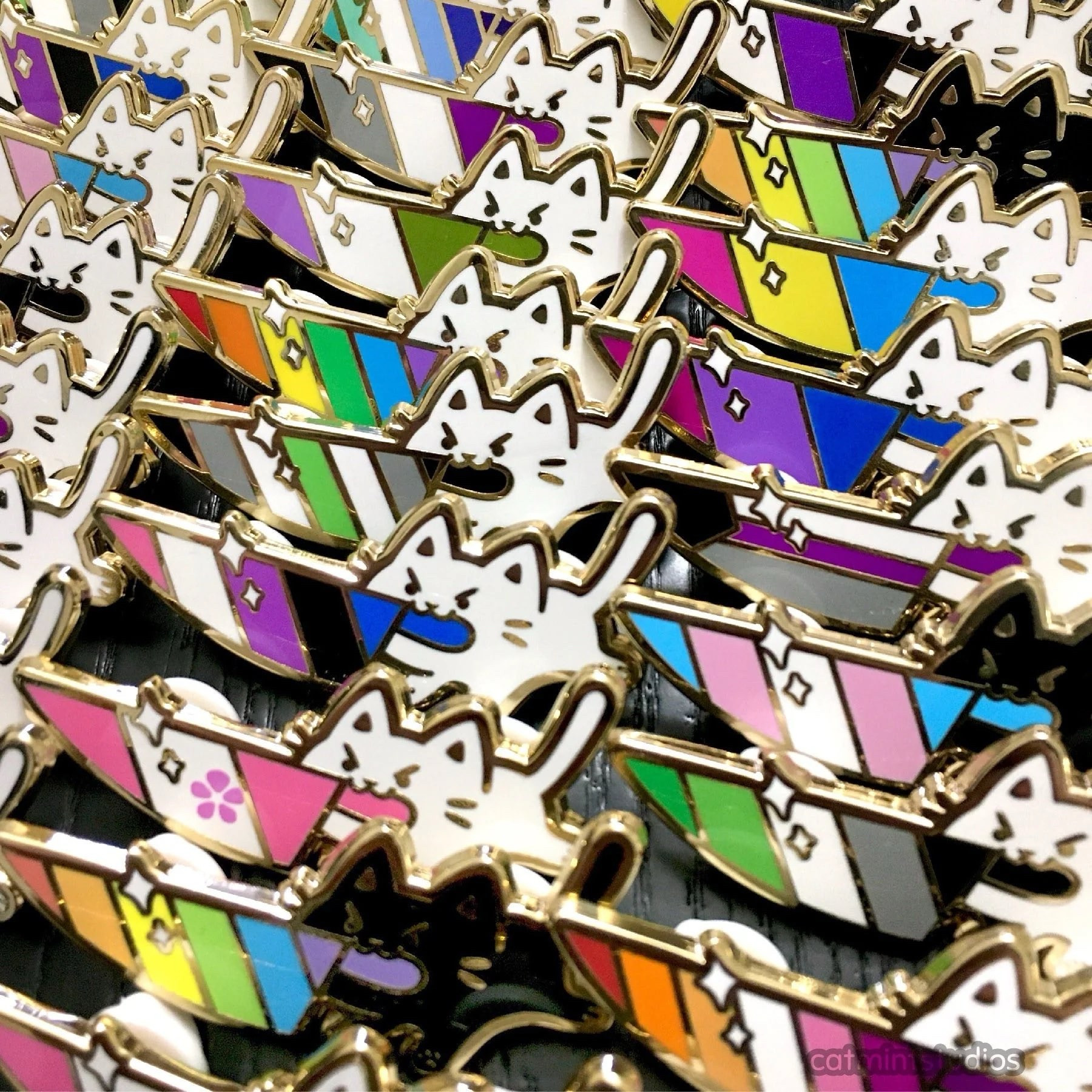 Different enamel pins are shown in rows. They are mostly white cats (and a few black cats) with angry eyes holding knives in their mouths. In the blades of the knives are different pride flag designs. 