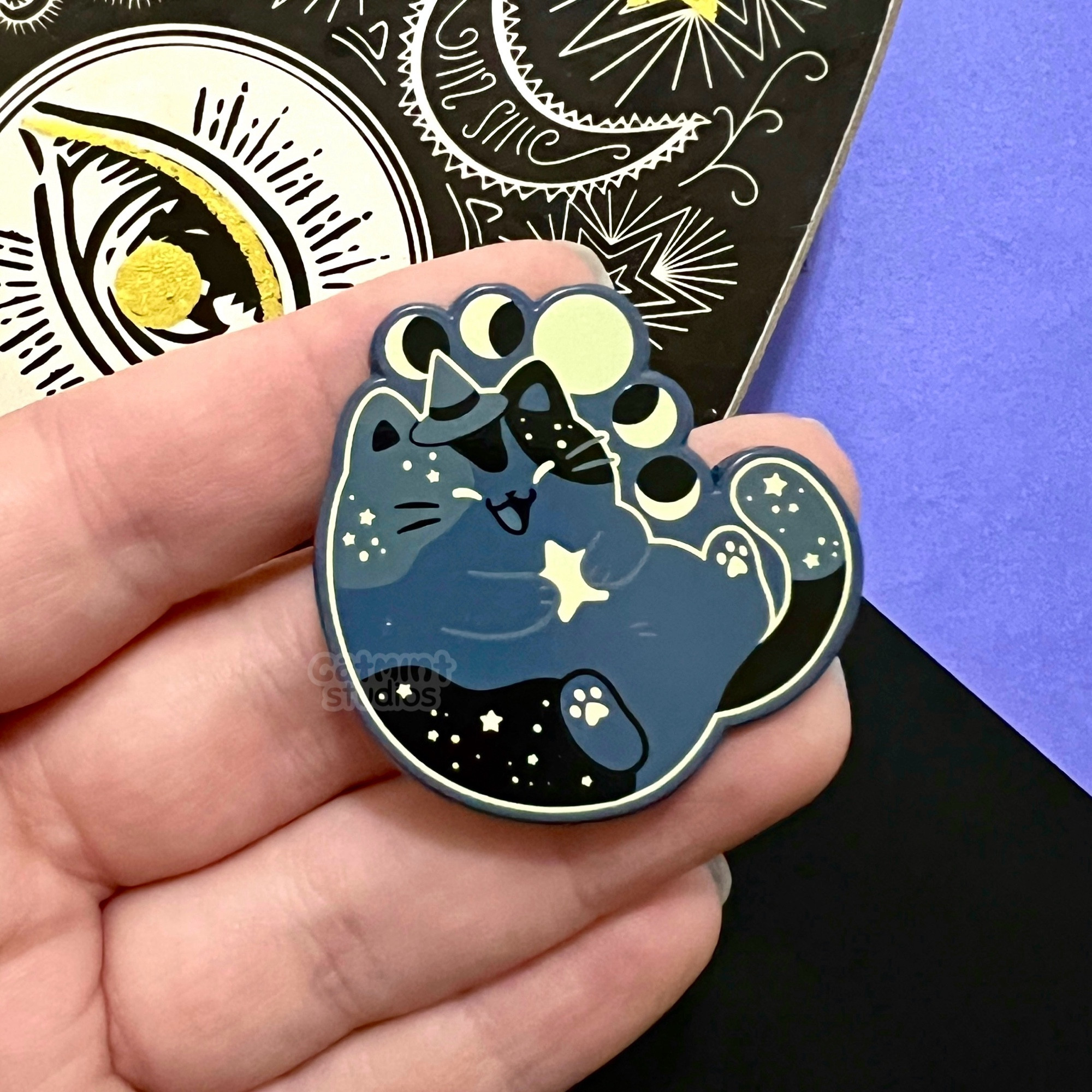 A hand holds an enamel pin on a black and purple background with a spooky planchette as a prop. The enamel pin design is of a chubby gray and black cat wearing a little witch hat and holding a star. There is an arc of moon phases above their head. There are little stars in the patches on their body. 