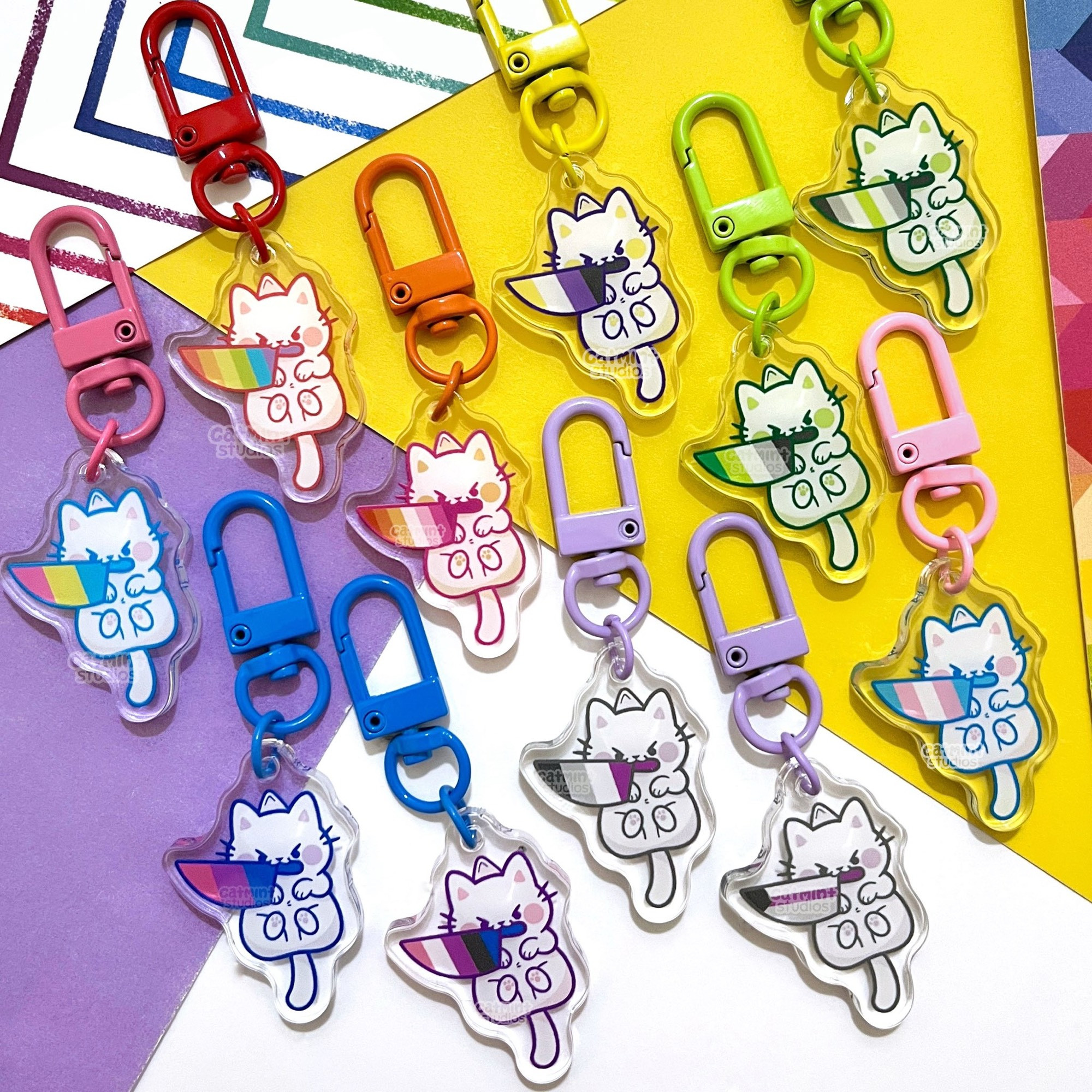 Various small keychains are shown. They are all little white cats with angry eyes holding knives in their mouths. In the blades of the knives are different pride flag designs. 