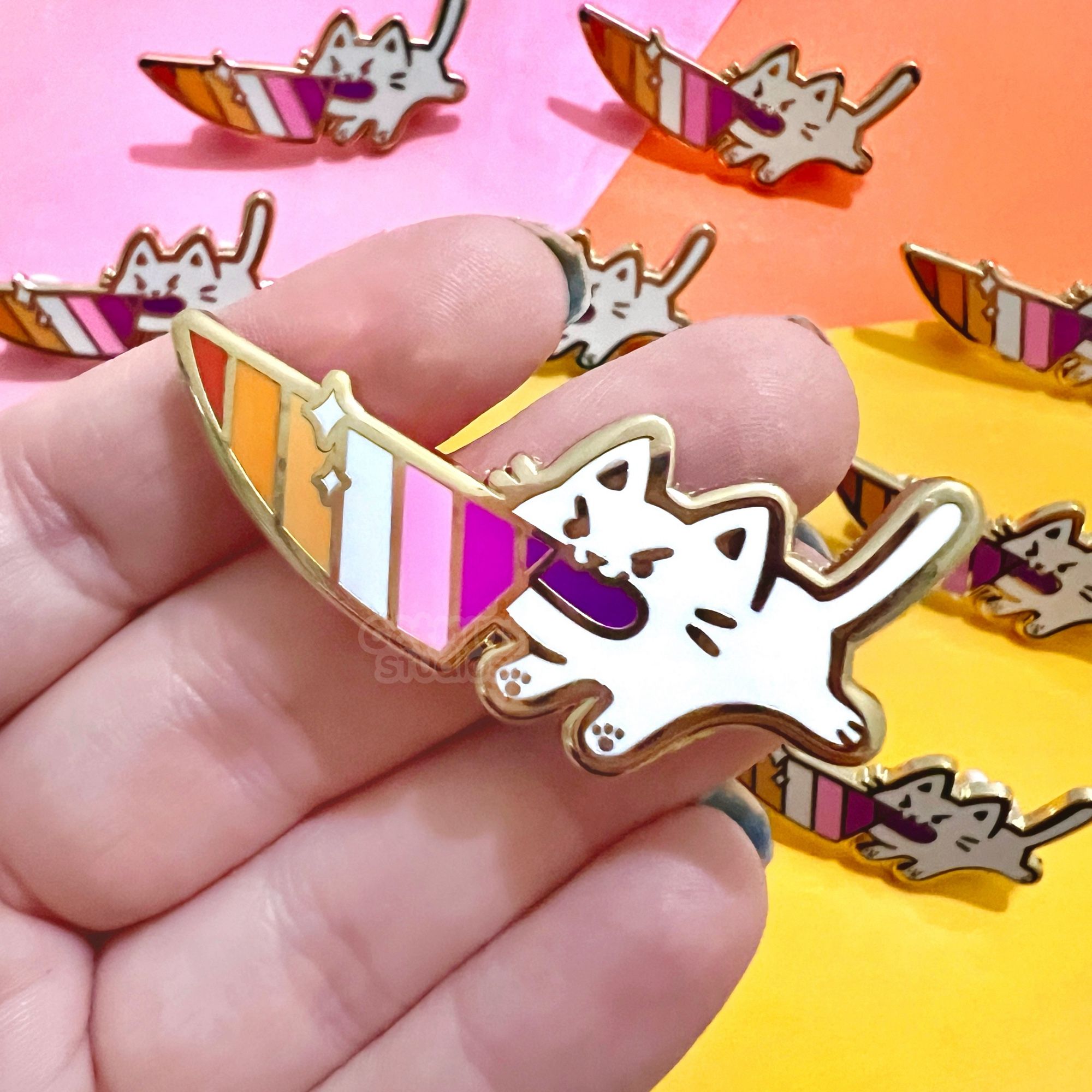 A hand holds an enamel pin above a pink, orange, and yellow background. Multiples of the same pin are also scattered in the background. The pin design is a small white cat holding a knife in its mouth that has the lesbian flag colors as stripes in the blade.