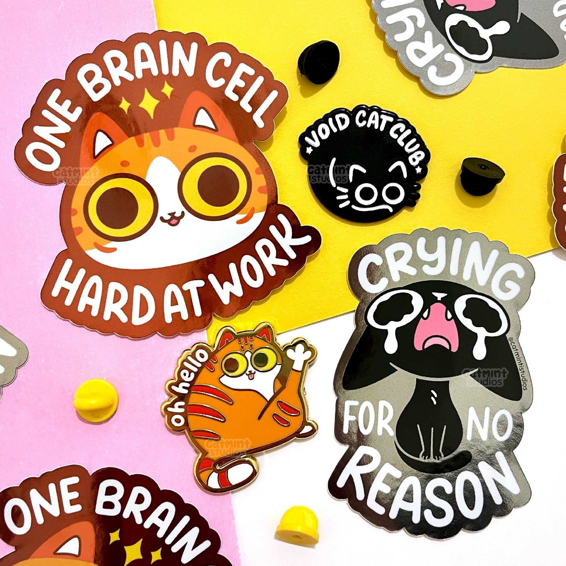 Stickers and pins sit on a pink, yellow and white background. There are 2 sticker designs - the first is a silly orange cat face with the tongue out and the words “one brain cell hard at work” ; the second is a crying black cat with the words “crying for no reason”. The first pin design is a chubby orange cat with his leg up and the words “oh hello.” The second is a curled up black cat with the words “void cat club”