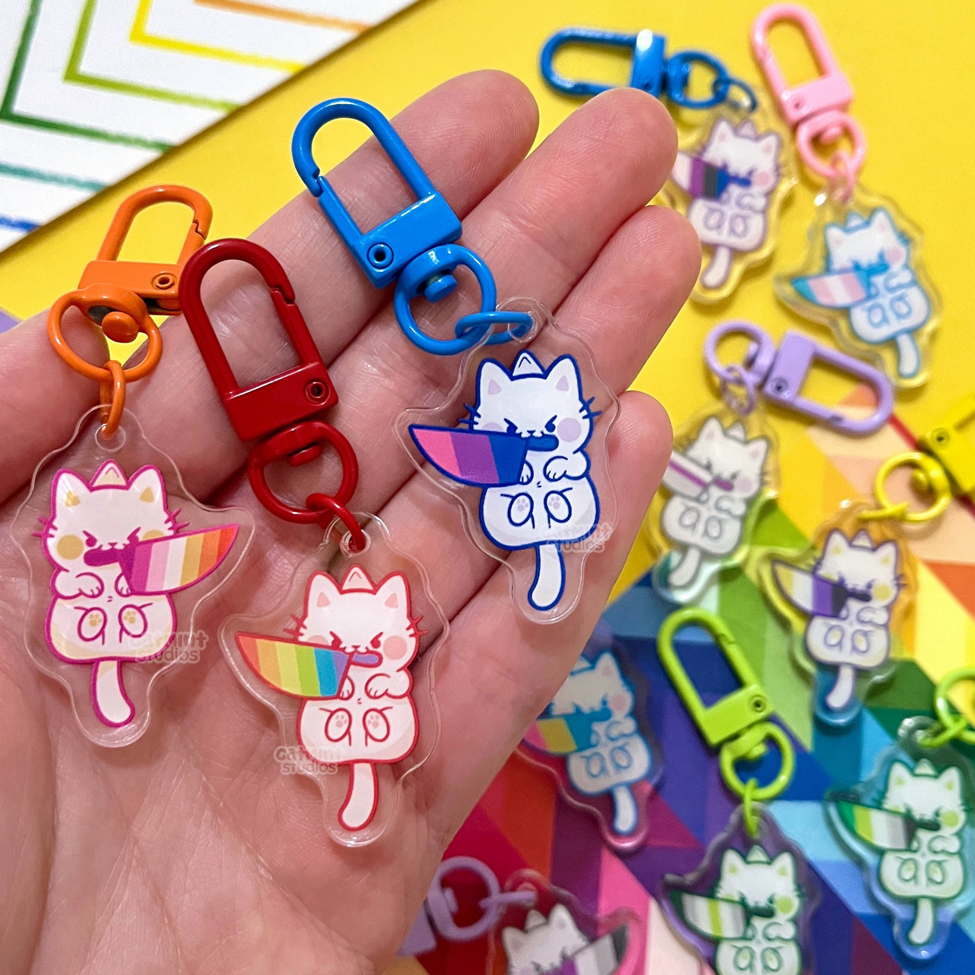 A hand holds three small charm keychains. The charm designs are small, cute white cats with angry eyes and knives in their mouths. The knives have the stripes of pride flags in the blades. In the hand are the lesbian, gay, and bisexual variants/flags. In the background are other charms with more different flags. All charms have a different color keychain clasp that corresponds to the flag colors.