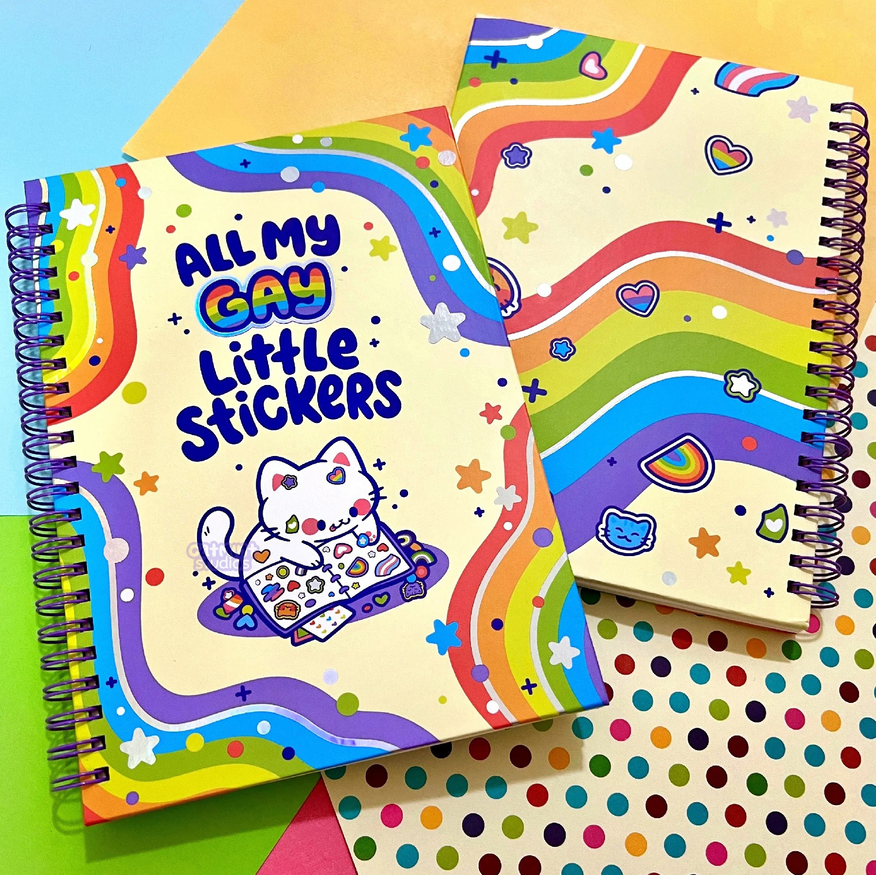 A sticker book is shown - the book has a purple coil. The front design has rainbow waves with stars and silver foil. It says “for all my gay little stickers” and has an illustration of a white cat putting stickers in a book. The back cover has more rainbow waves and little sticker designs. 