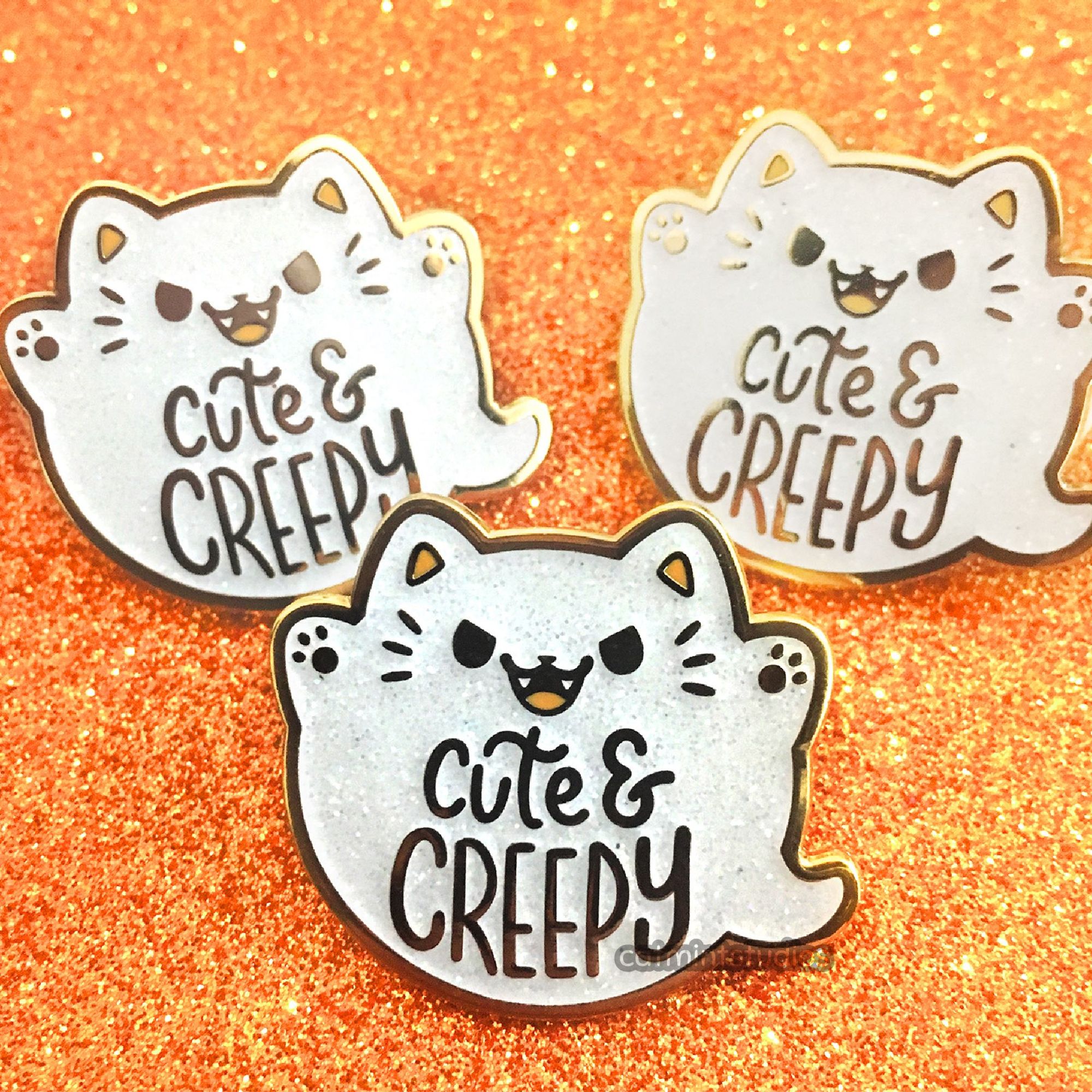 Three enamel pins of very round ghost kitties. They are glittery white, and have angry cute smiling faces with open mouths. Their tiny paws are raised and on them it says “Cute & Creepy.”