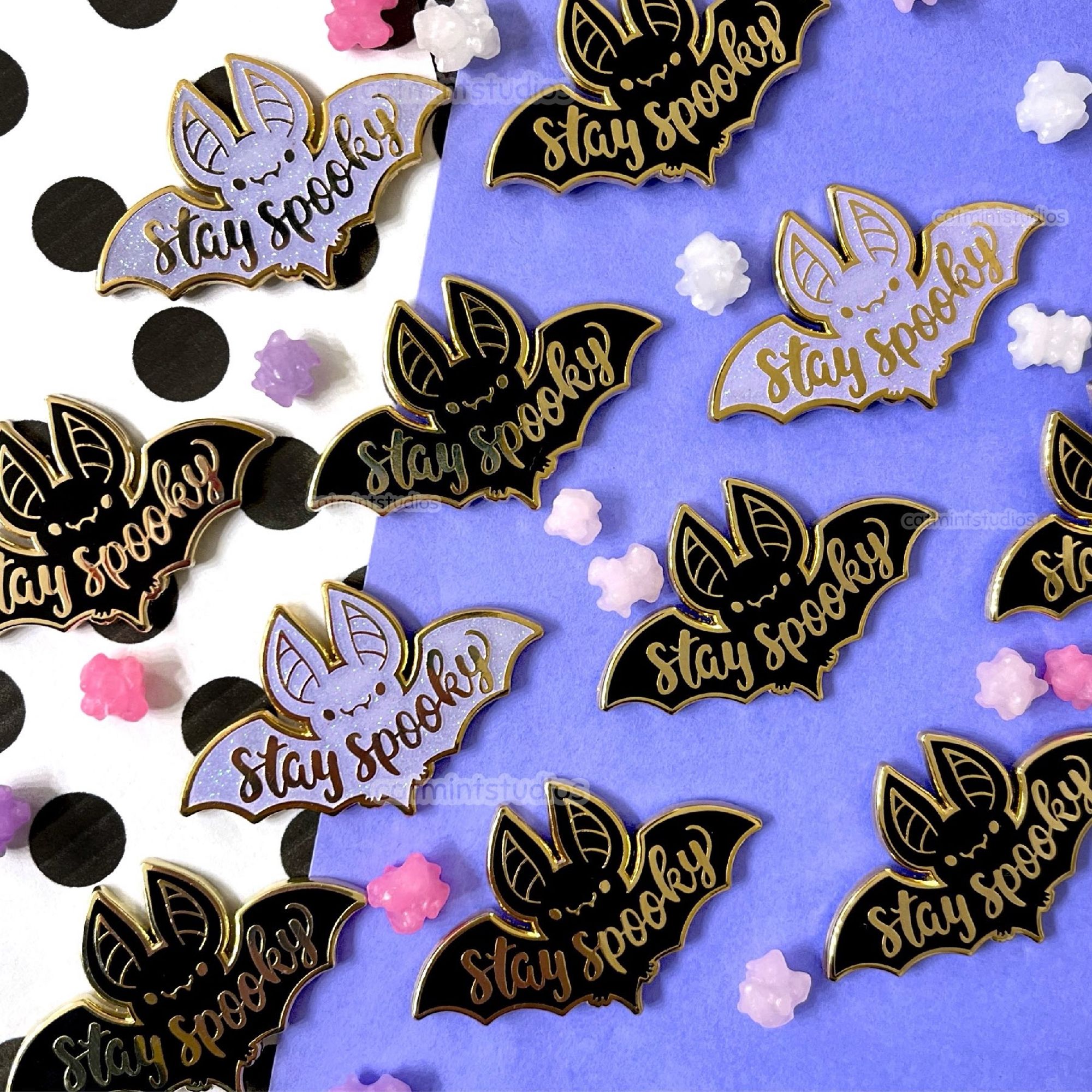 Enamel pins are laid in rows on a black, white, and purple background with konpeito candies scattered around. The pin is a bat design that says “stay spooky” in script across the body. The pins come in black and lavender.