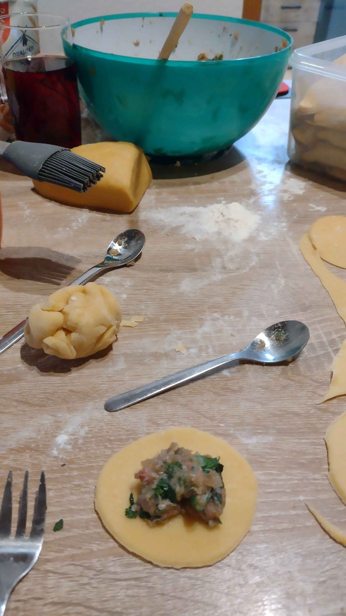 A photo showing a kitchen table full of utensils for making german ravioli. On top you can see a blue bowl with meat and spinache filling, on the side you have dough and a few spoons and forks. In the lower halfe there is an unfinished ravioli circle with filling that is about to be folded