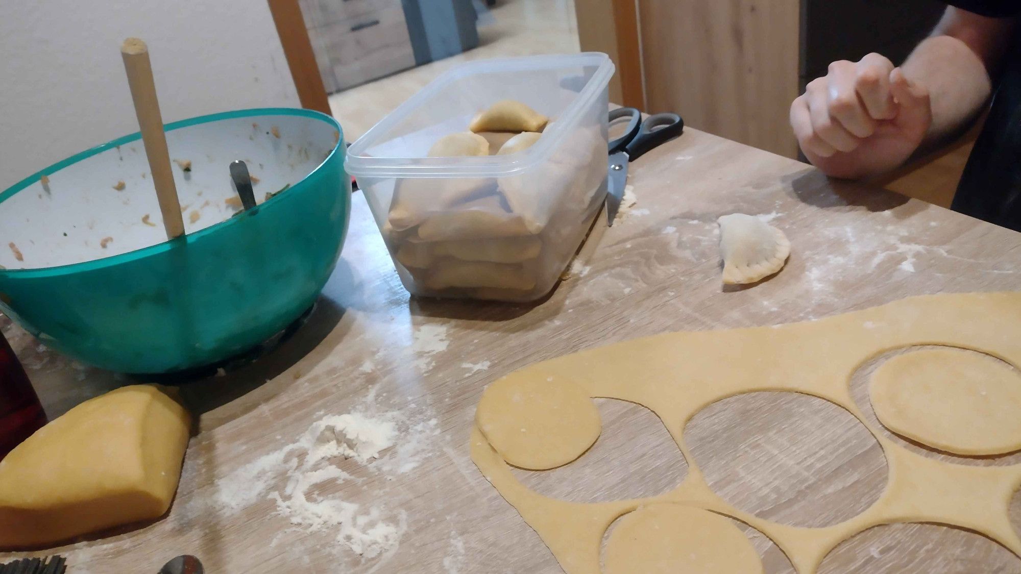 A photo showing a kitchen table full of utensils for making german ravioli. On the left you can see a blue bowl with meat and spinache filling, on the side you have dough and a few spoons and forks. You can see dough thats already flattened and seperated into little dough circles for further german ravioli production