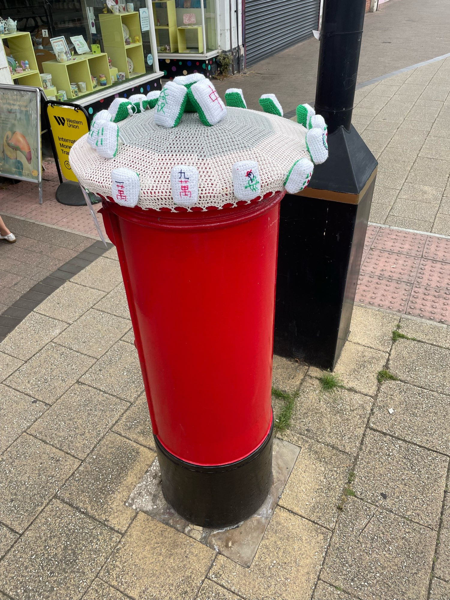 A photo of a red U.K. post box with a knitted hat like cover on the top. The cover has 3 mahjong pieces sewn in the centre and many sewn around the edges. The post box is in the middle of a walkway and a lamp post and a some store fronts are visible in the background.