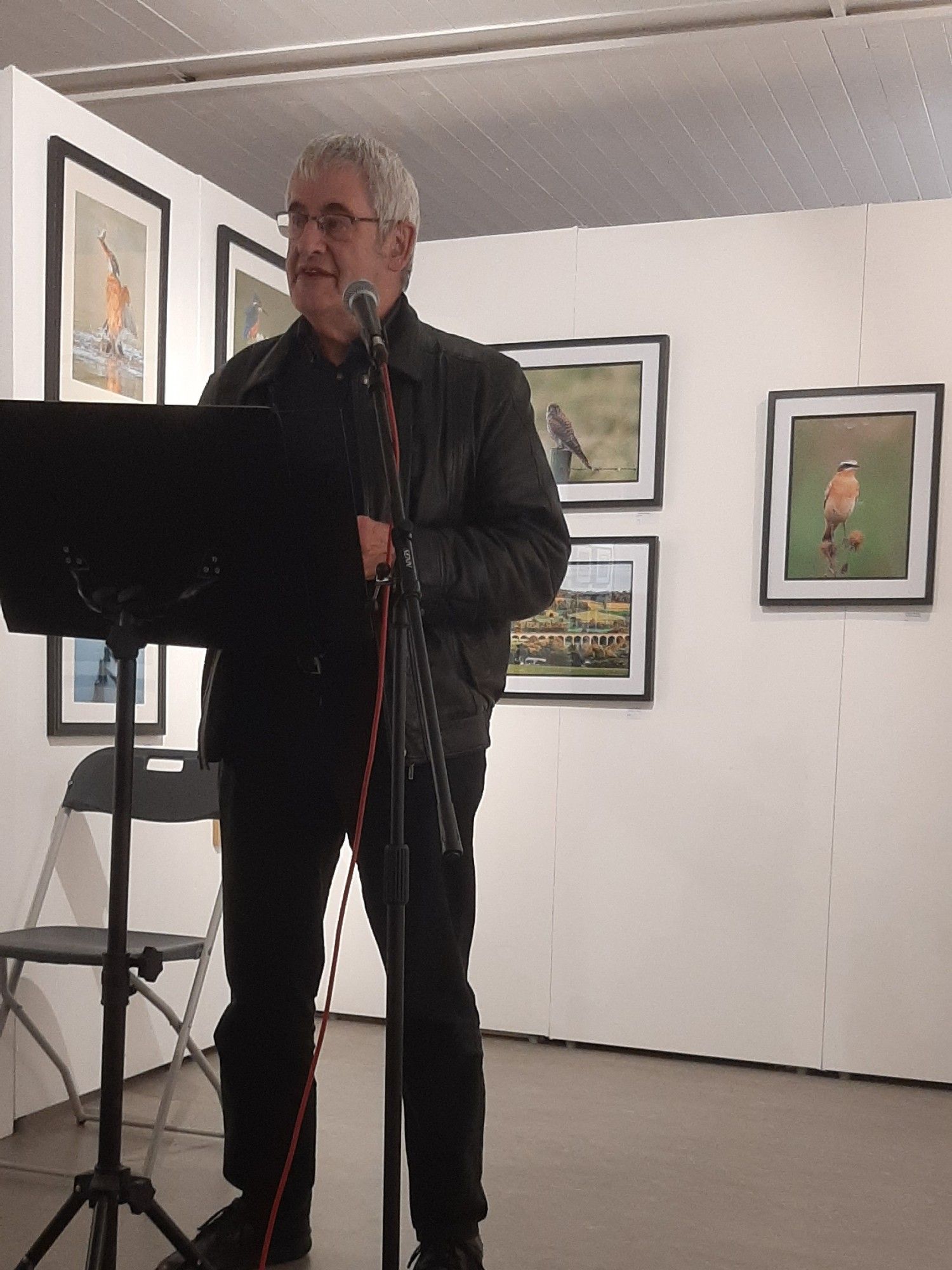 Photo of Ian Clarke dressed in black behind a microphone in an art gallery with photos of wildlife on the white wall behind him.