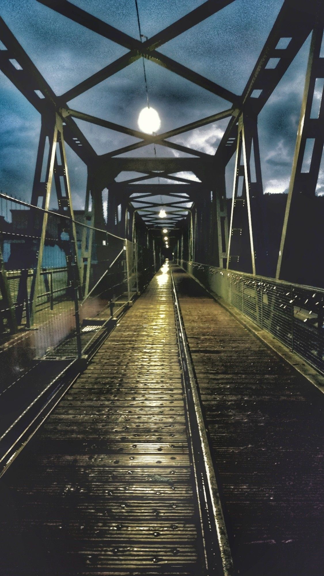 Blick auf eine Brücke aus Stahlträgern, den Schienen folgend, die sich bis in die Mitte des Bildes ziehen. Lins, rechts und oben Stahlträger mit weißen Hängelampen. Das Licht spiegelt sich im regennassen Holzboden. Der Himmel ist dunkel bewölkt.