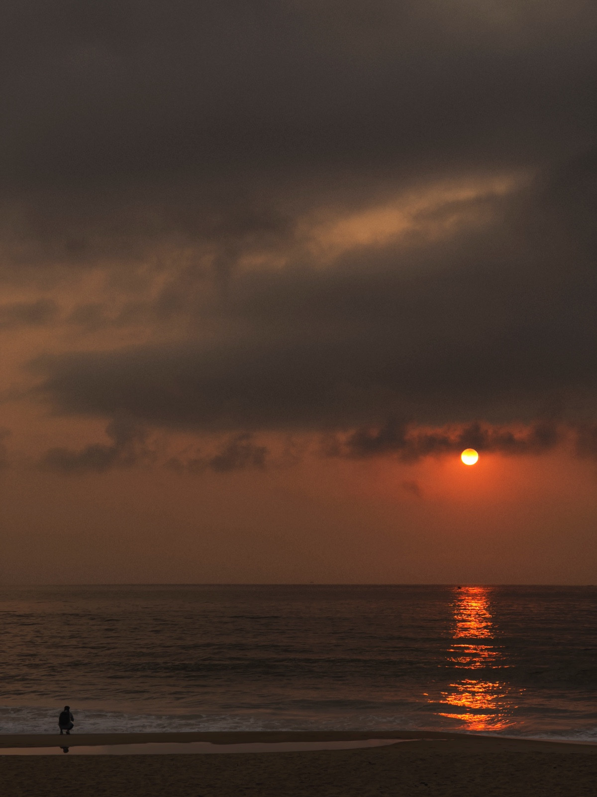 A post thunderstorm sunrise at the beach with dark thunderclouds high above. A deep orange sun is well above the horizon as it casts a beam of light on the water. A lone man is on his haunches taking in the scene.