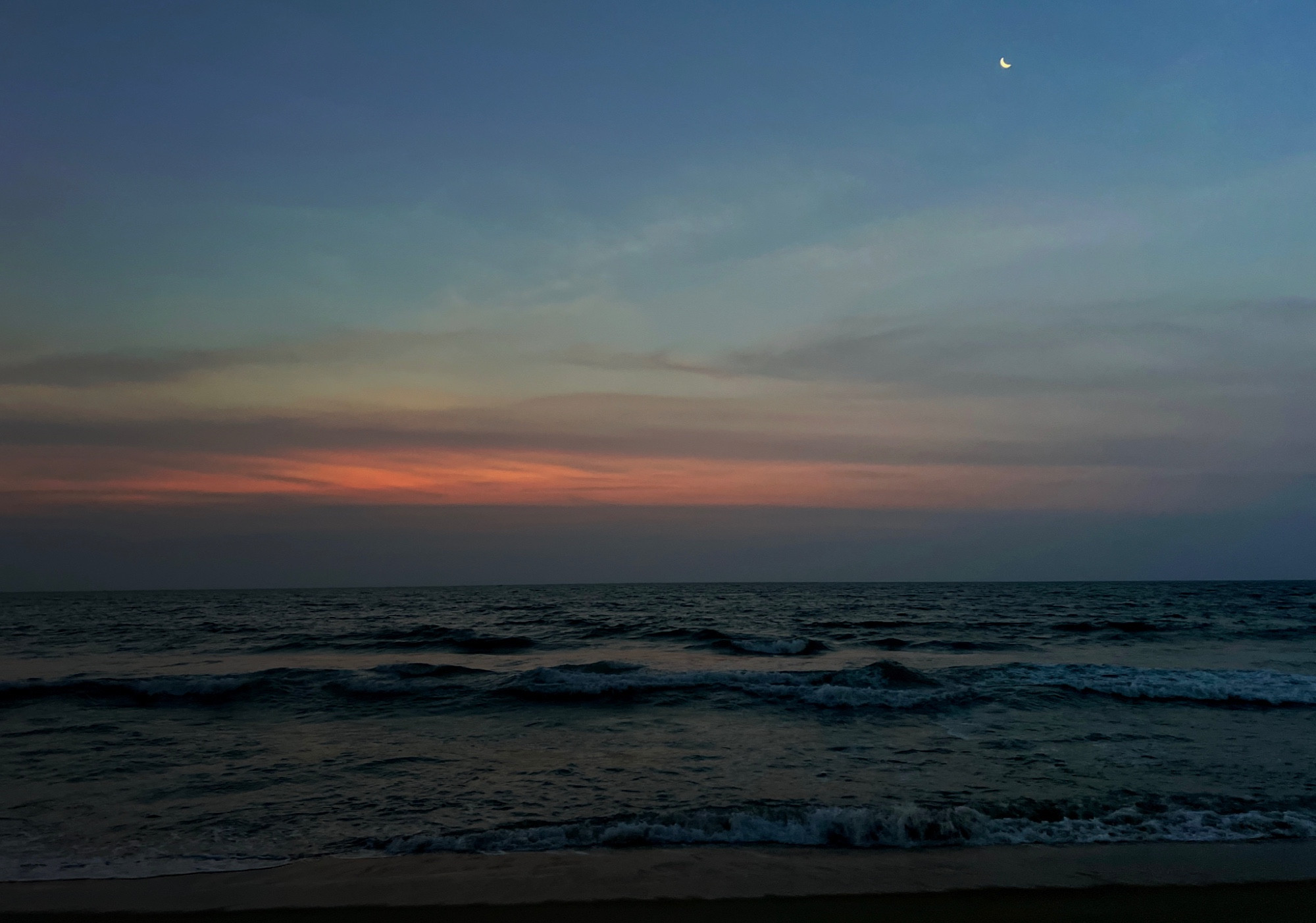 A bluish sunrise at the beach. The sun above the horizon but light is diffused by long thunderclouds. A crescent moon is also seen high above the horizon towards the top right.