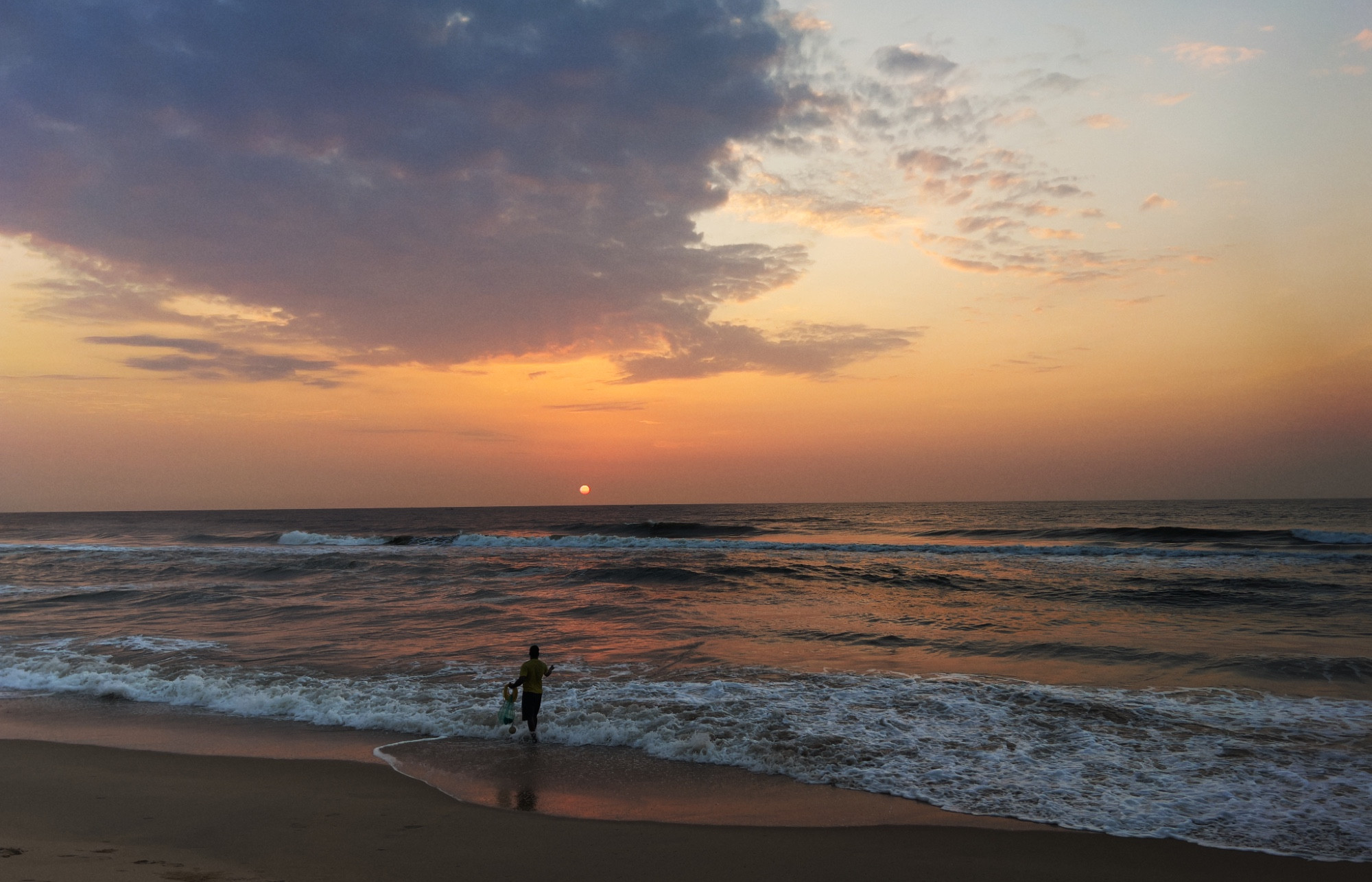 A dramatic sunrise at the beach with orange, pink and bluish grey hues. The sun is a small dot just above the horizon. A lone fisherman in the foreground is on the beach trying to throw his net as waves wash over.