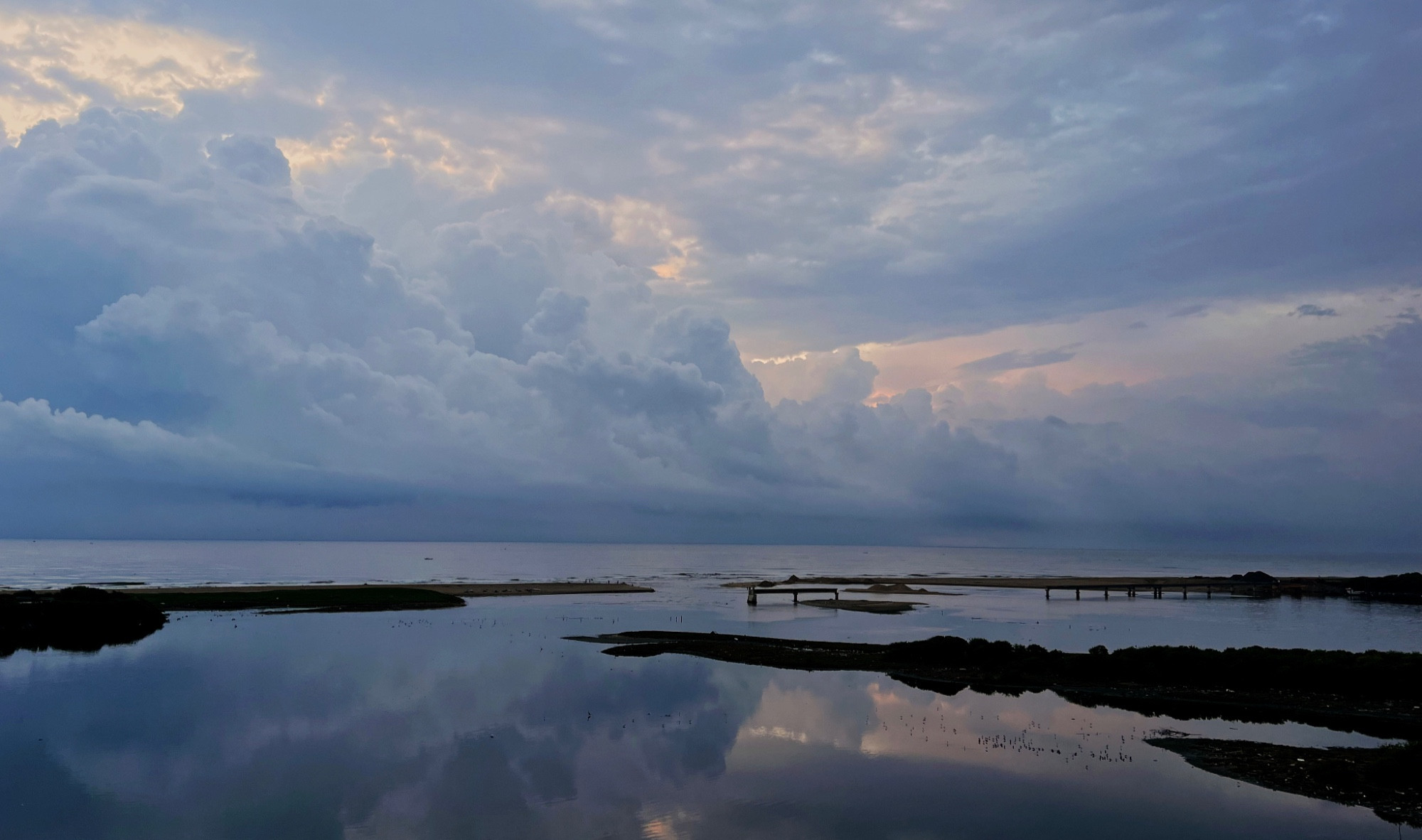 Thick clouds obscure a rising sun making for a dramatic scene in the sky. In the foreground is a calm sea with sand bars and remnants of a half finished bridge.