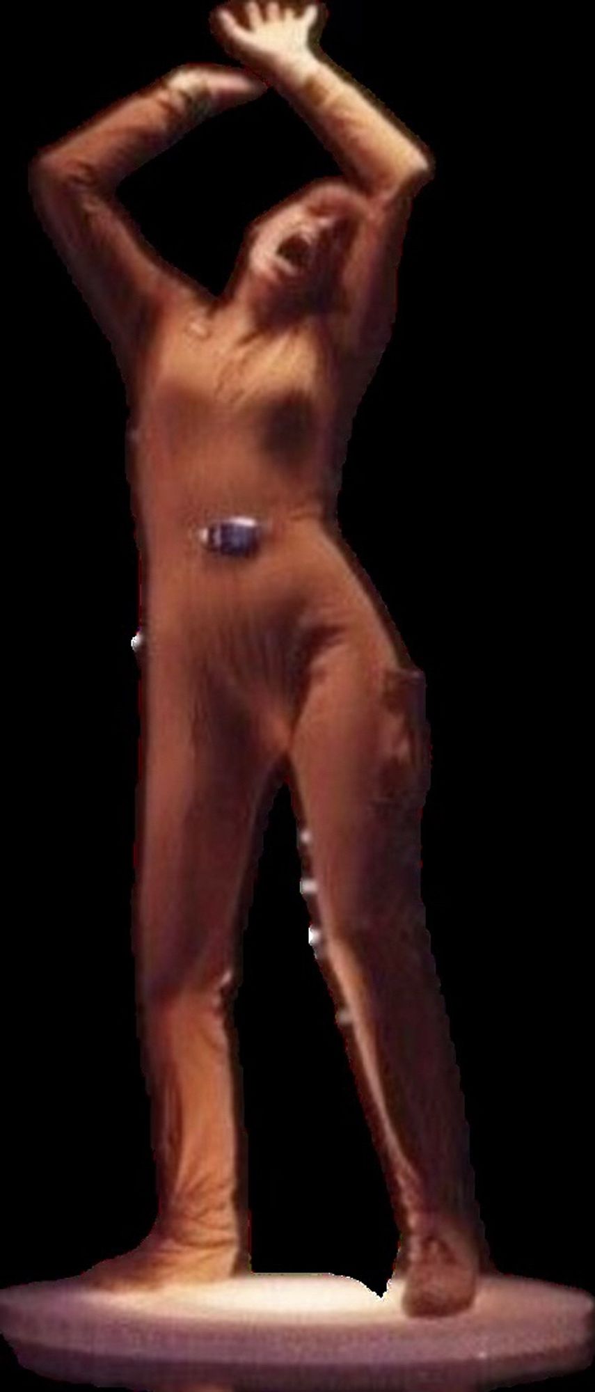 A woman standing, legs apart, hips askew, arms raised far above her head in a protective manner, her head tilts into back and to the left with mouth stretched open.
She wears a medium brown jumpsuit with a small black belt buckle.
It’s hard to say if she’s yawning, screaming in agony, listening to funky music or working through the burn of exercise.