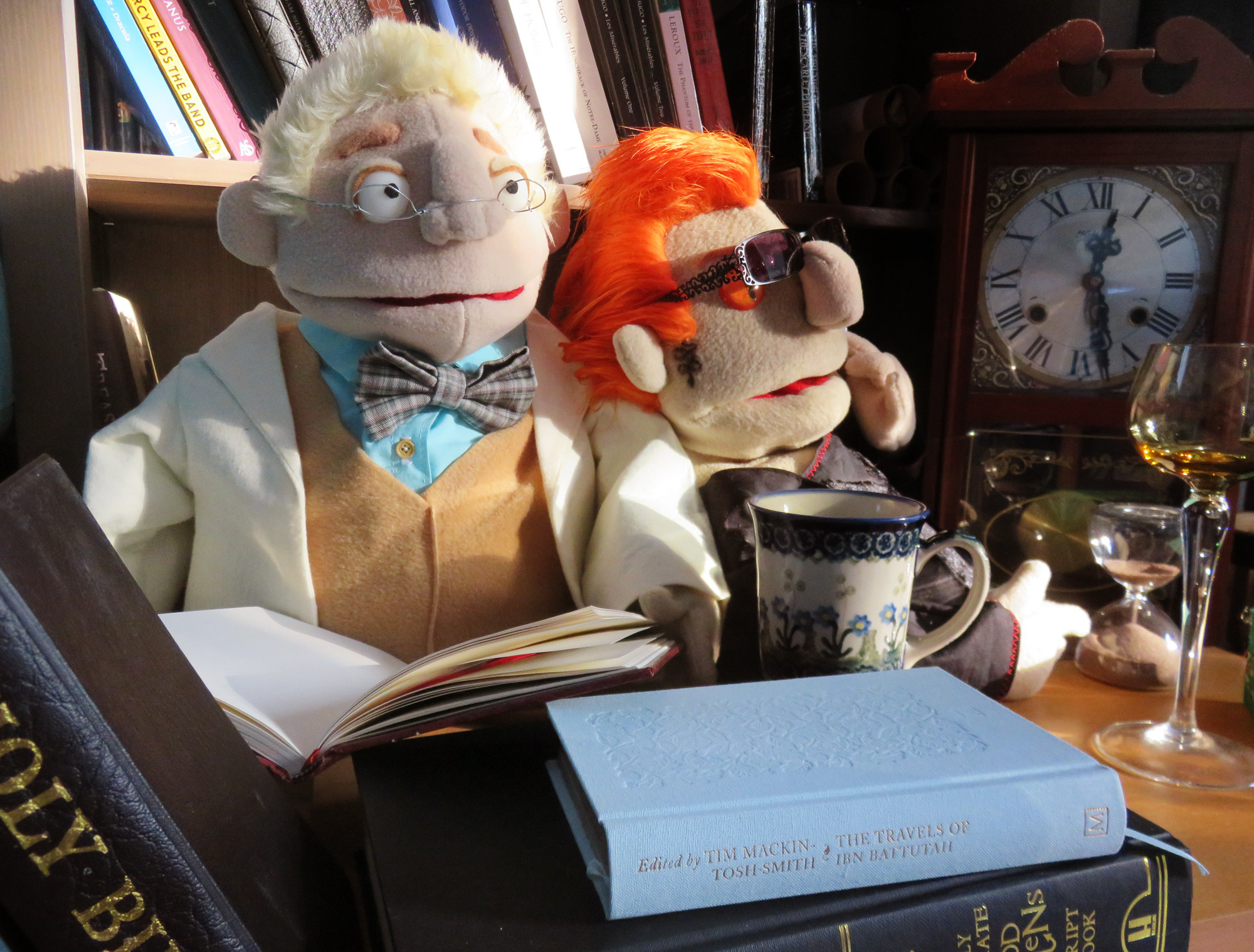 A photograph of muppet version of Crowley and Aziraphale from Good Omens arranged in front of a bookshelf and grandfather clock, surrounded by books with a cup of tea and wine glass on the surface in front of them