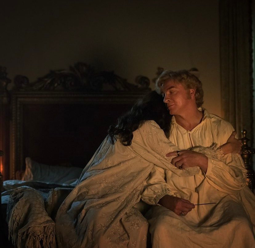 A shot from episode 10 of season one of our Flag means death. Sitting on the end of their bed by candlelight, Mary Bonnet embraces Stede Bonnet. Both of them are in their nightgowns.