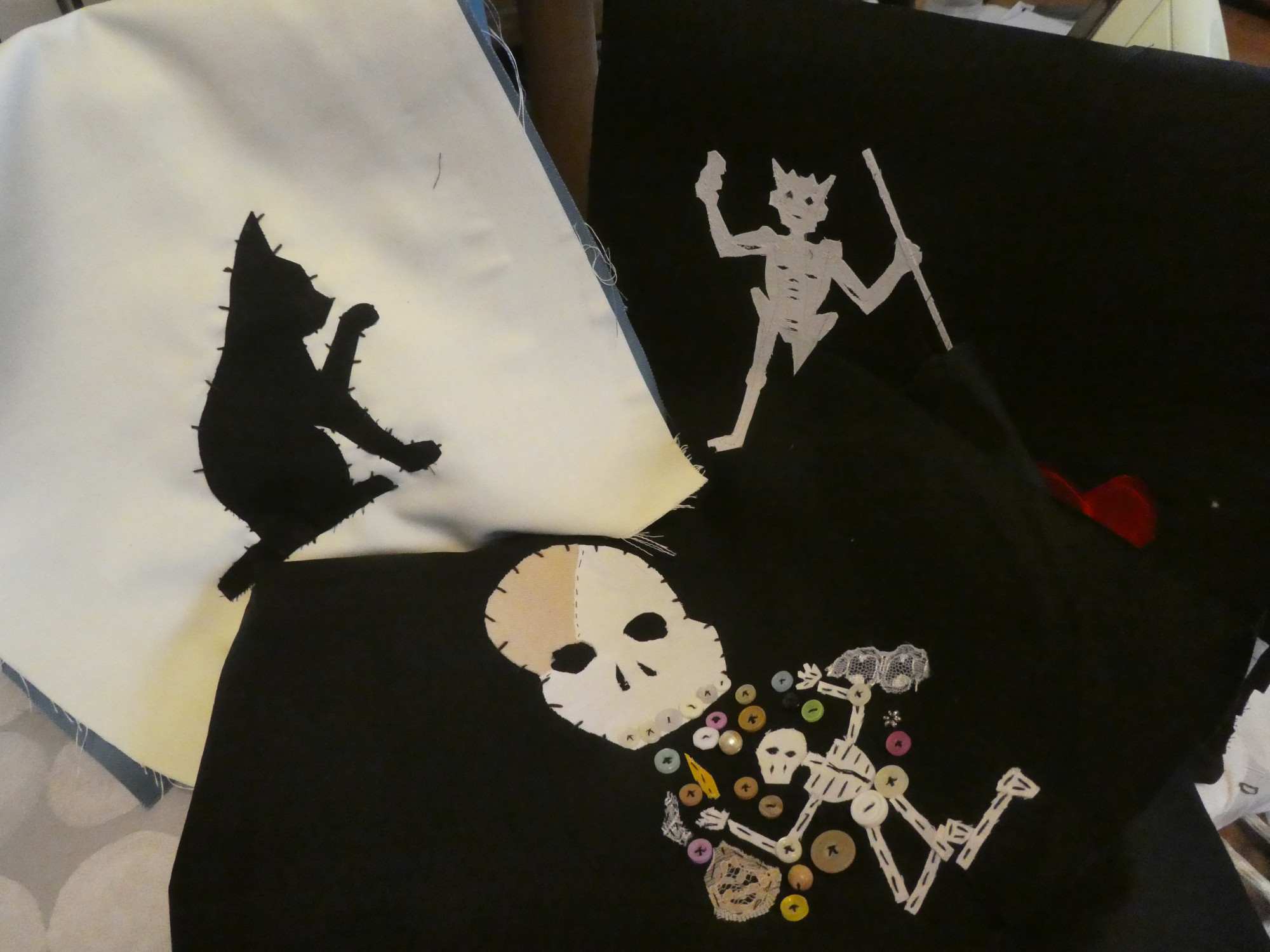 a photograph of three flags from Our Flag Means death, handstitched and rendered on bags. One is white with a black cat stitched on, the other two are both black, one with a horned skeleton stabbing a heart and the other with a large skull vomiting buttons on a small skeleton