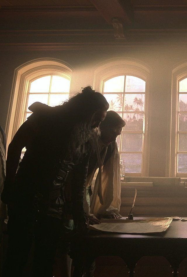 A still from episode 9 of season 1 of our flag means death. Blackbeard and Stede Bonnet are looking down at a parchment on the table in front of them, lit from behind by golden sunlight coming through the windows of the ship's cabin.