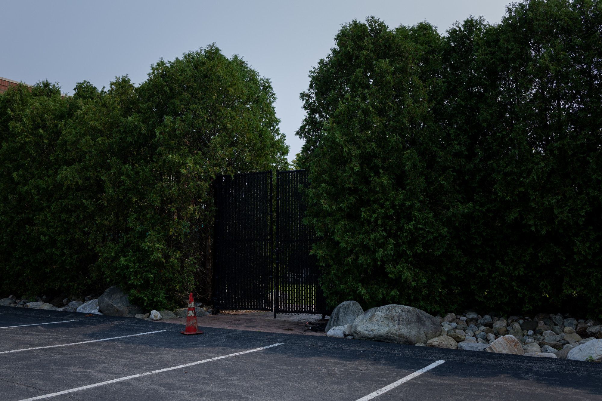 A wall of hedges, at the edge of a parking lot, with a black lattice gate in the middle. An old weathered pylon sits in front of it.