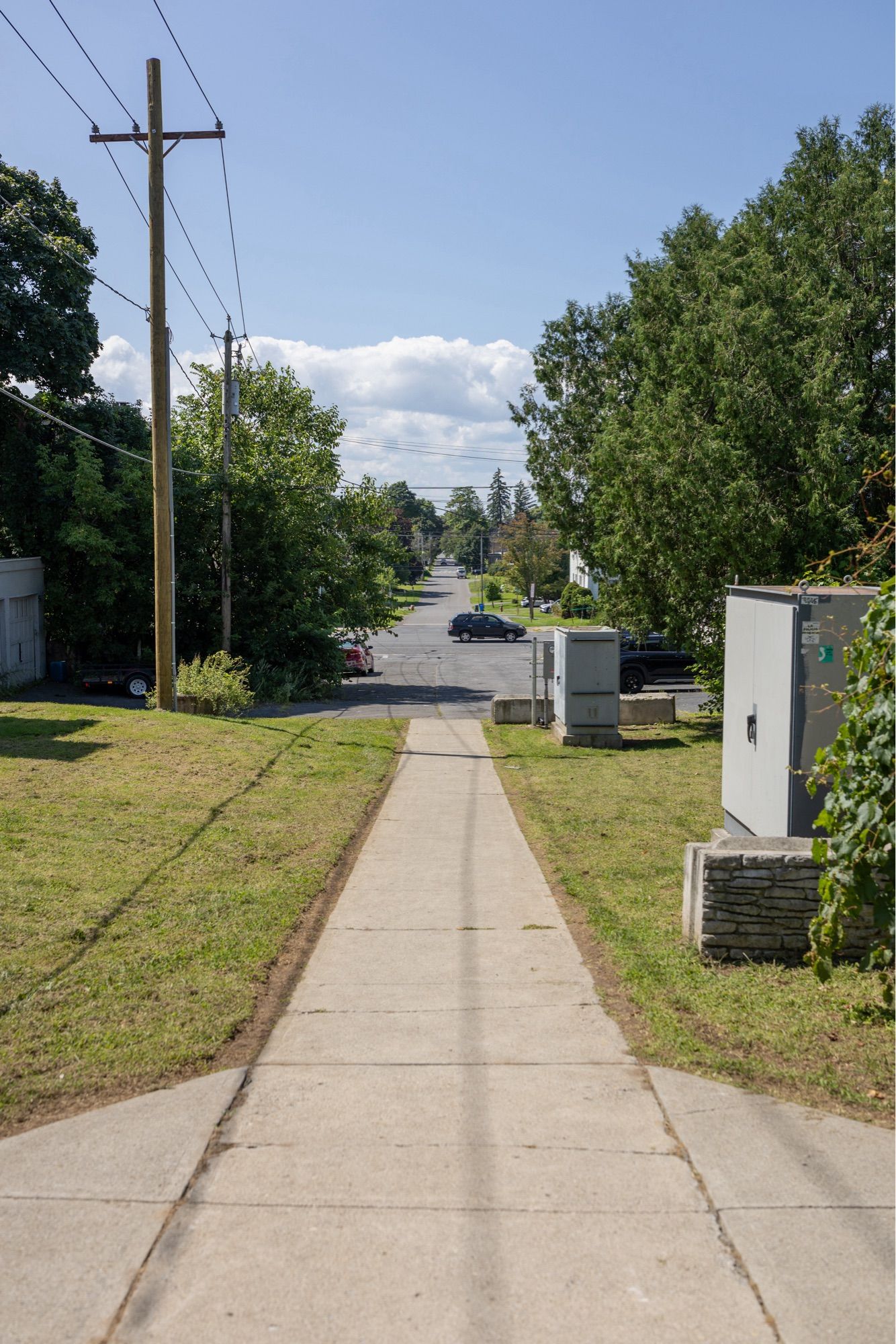 Photo of a sidewalk that leads directly into a road which goes even further. Two gray electrical boxes are on the side.