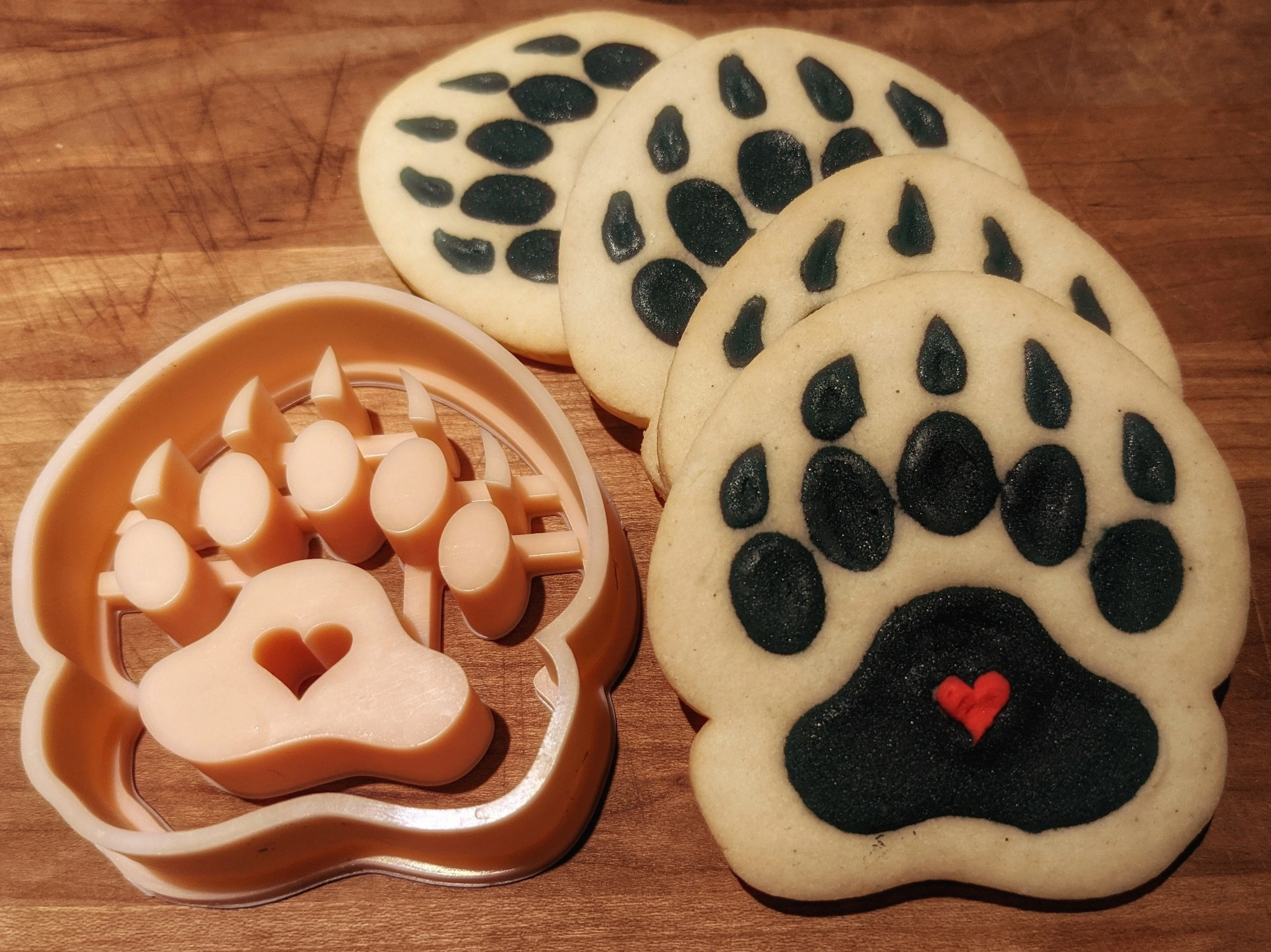 On the left is a beige cookie cutter, it has the outline of a bear paw print with a heart in the middle. To the right are four sugar cookies fanned out. They're golden brown with black royal icing forming the shape of the paw and a little red icing heart in the middle.