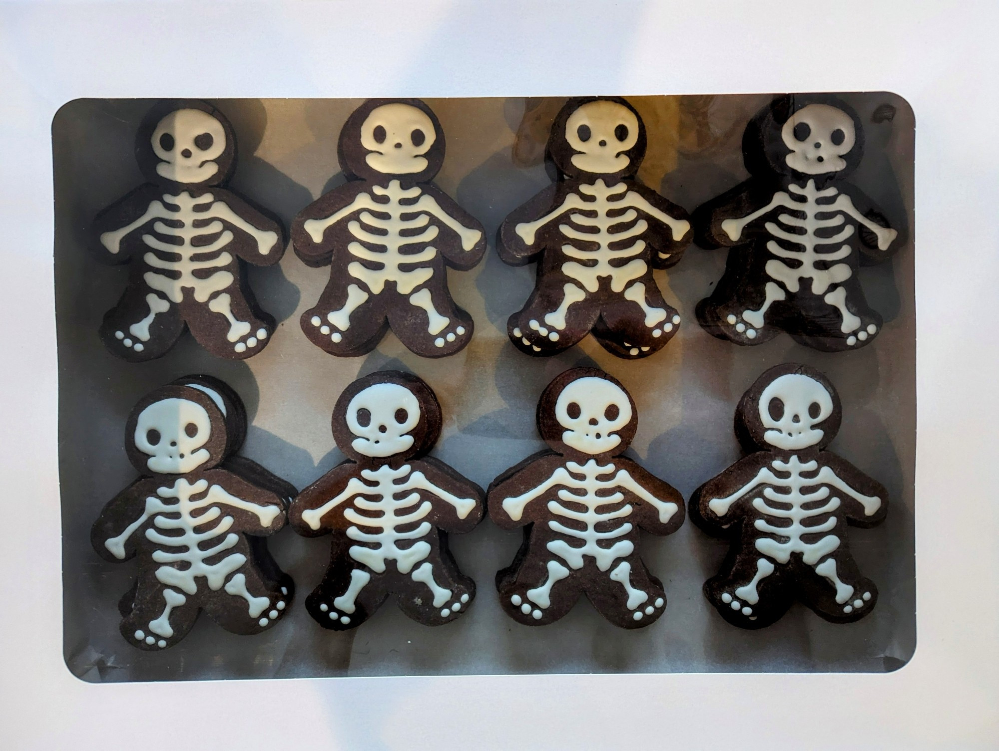 A box of cookies, arranged in two rows. The cookies are chocolate brown and have white icing creating the bones of a skeleton. Their jaw makes them look like they're smiling and they have three little toes on each foot, they're funny not scary cookies.