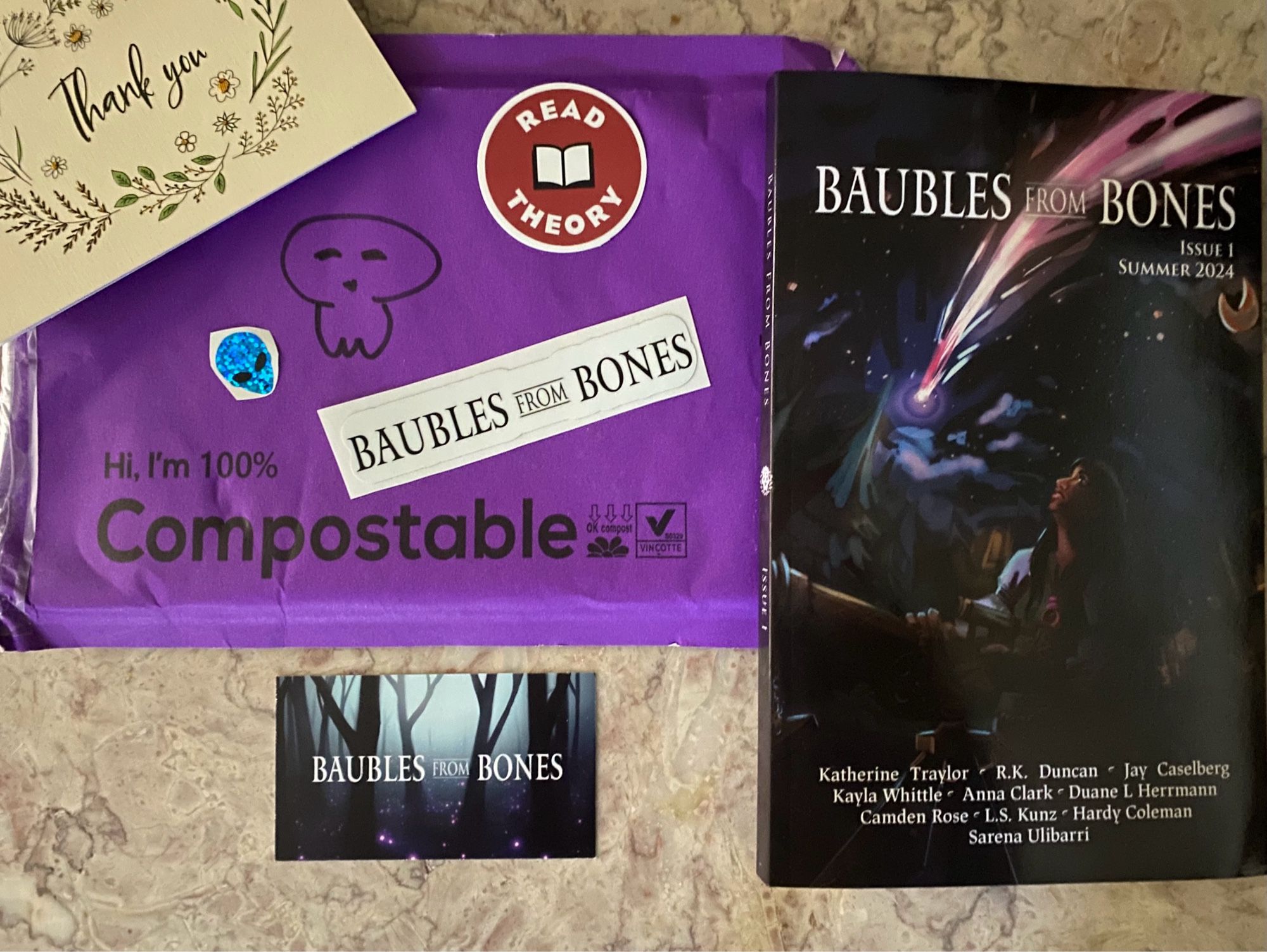 Book (Baubles From Bones) with a collection of alien and reading stickers next to a thank you card and purple packaging.