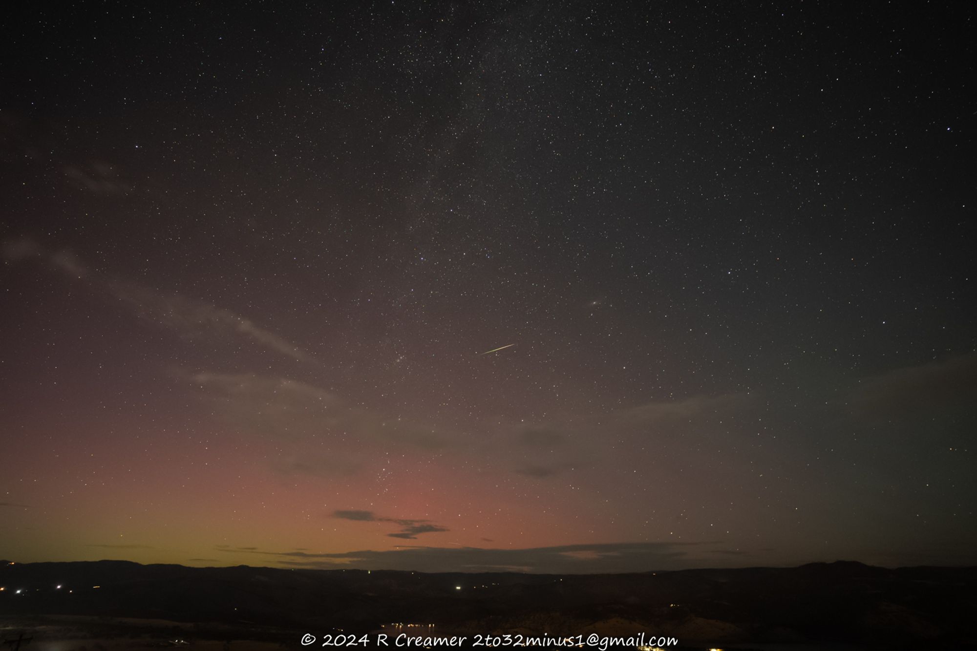 Photo of night sky showing one tiny Perseids shower meteor streak + a tiny/fuzzy Andromeda galaxy (M31) just above and to the right of the streak + yellow-to-red aurora borealis lighting on the horizon, all with central California foothills at the bottom of the frame.