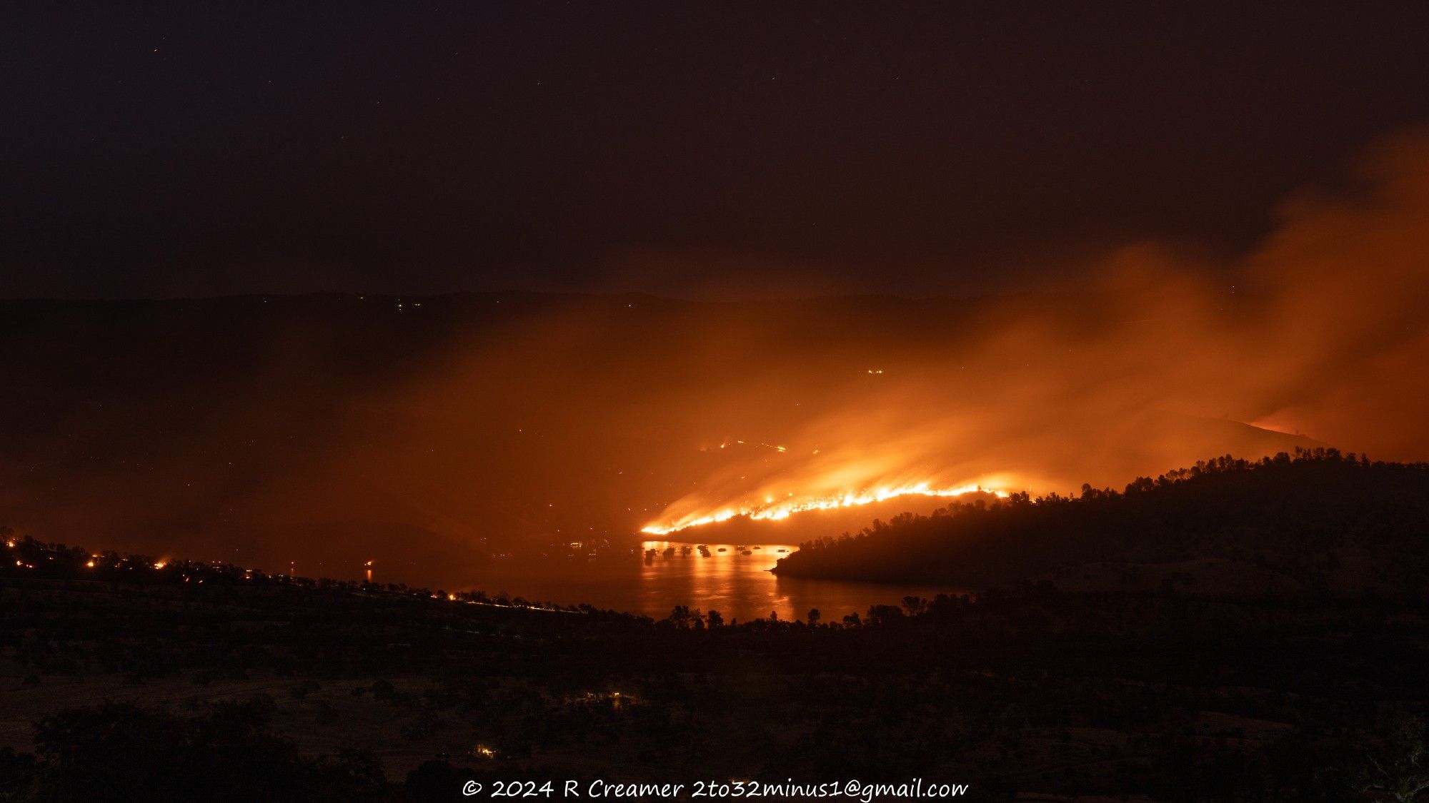 A view of Lake McClure's Barrett Cove with moored houseboats and behind them burning hills and glowing smoke