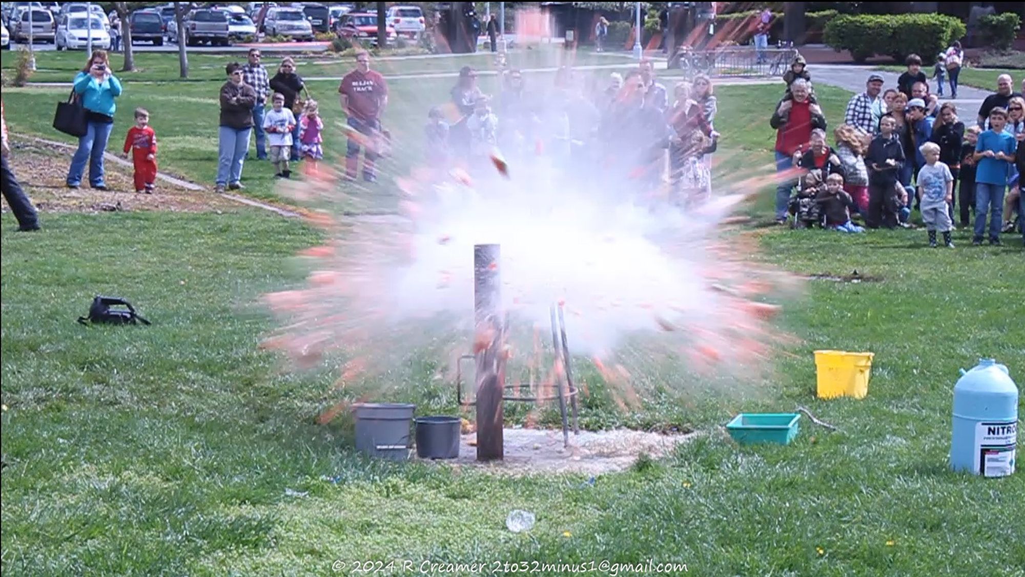 A photo of an exploding watermelon with pieces flying off in all directions at a high speed. Setting: in a field of grass with spectators observing at a safe distance.