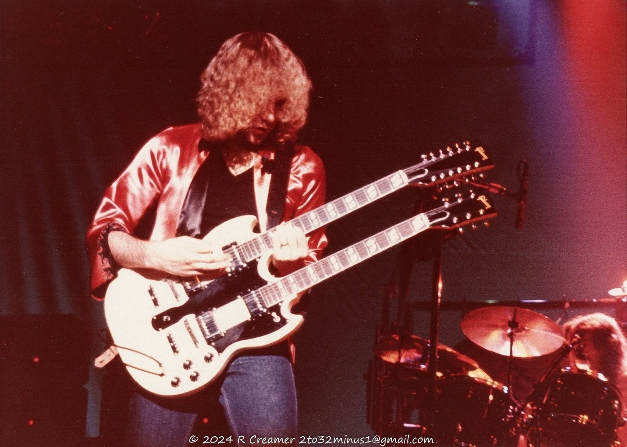 A photo of Alex Lifeson on stage playing a white Gibson double-neck (6/12 string) solid-body guitar with Neil Peart (drummer) visible in the lower-right corner with his drums/cymbals.