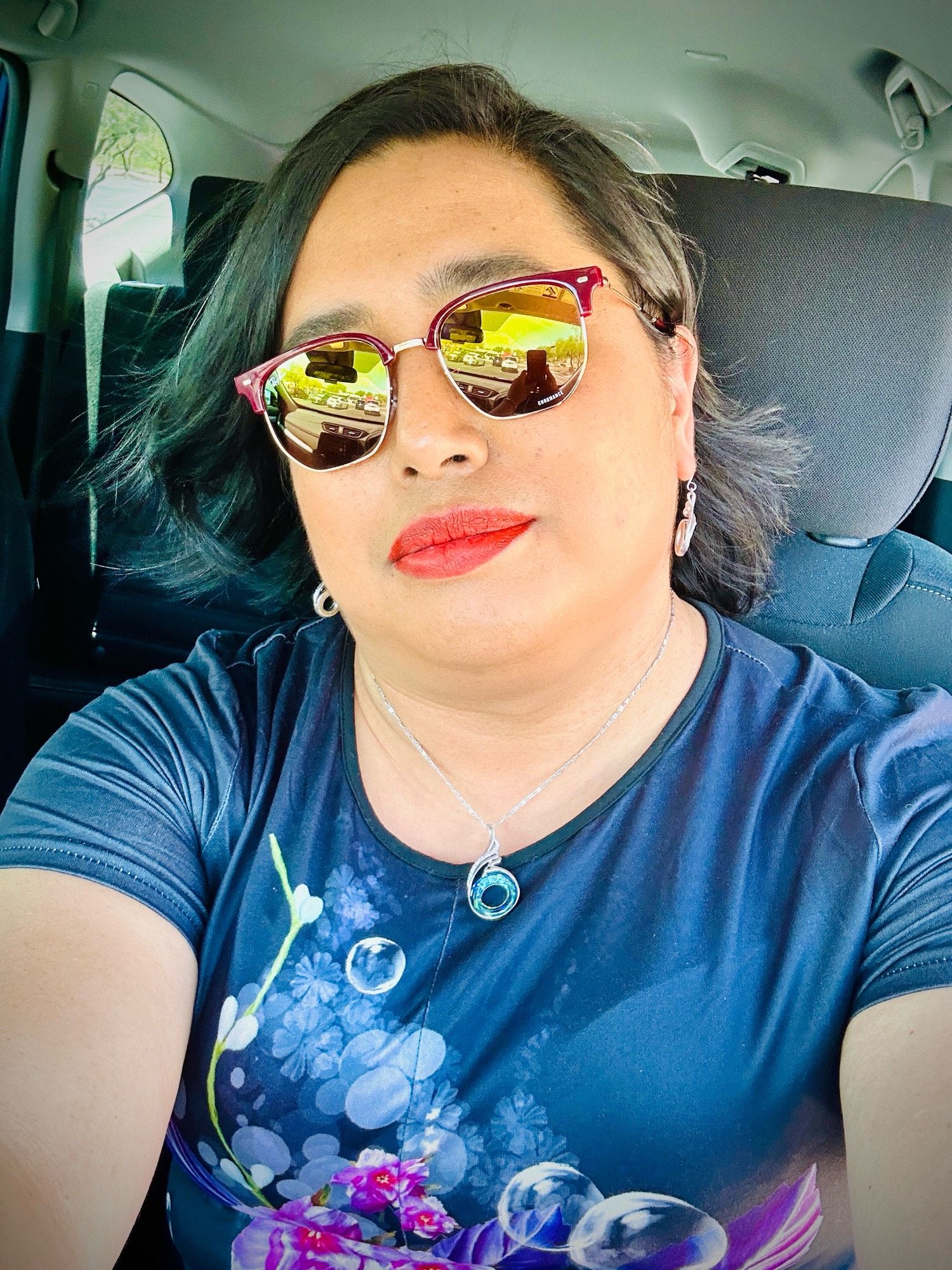 Trans AAPI woman in car wearing dark top with flowers and bubbles printed on it, a circular necklace and earrings, red lipstick and red sunglasses with shoulder length black hair blowing from the AC