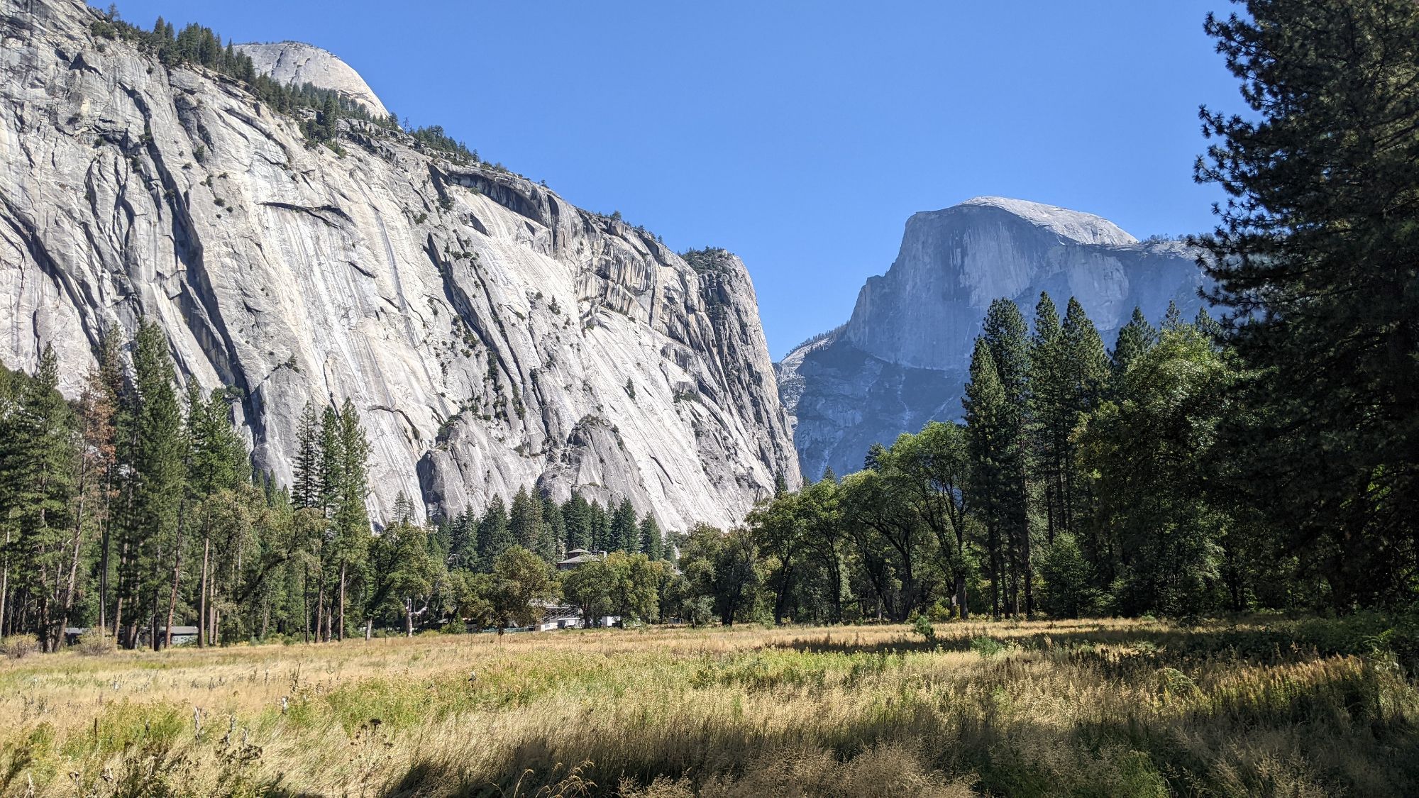 Granite cliffs tower above a dry, brown-green meadow in Ahwahne (Yosemite Valley). North Dome is peeking over the left cliff, and Tis-sa-ack (Half Dome) is on the right side.